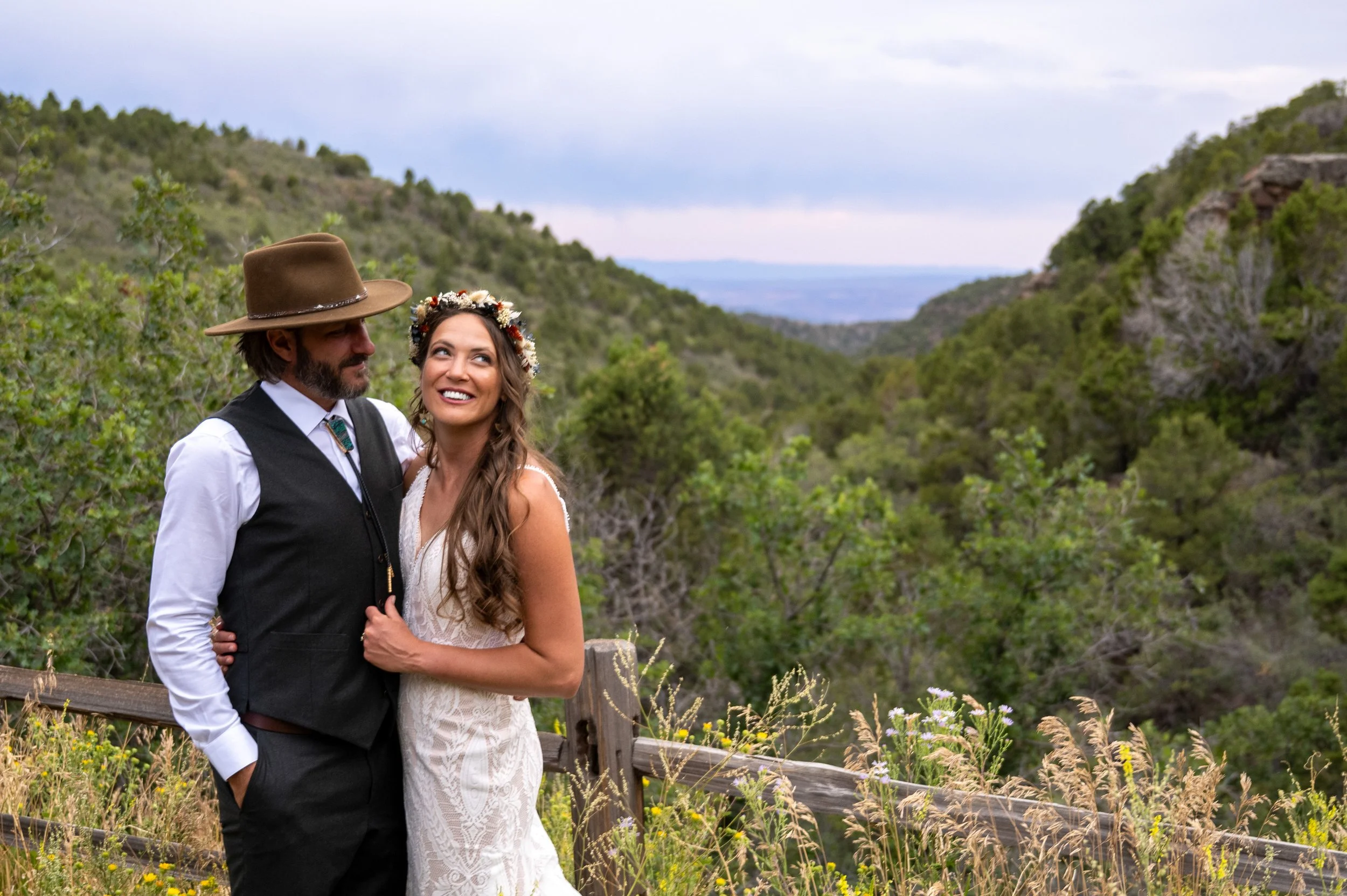 A smiling bride and groom in wedding attire standing outdoors on a mountain trail with lush green trees and hills in the background.