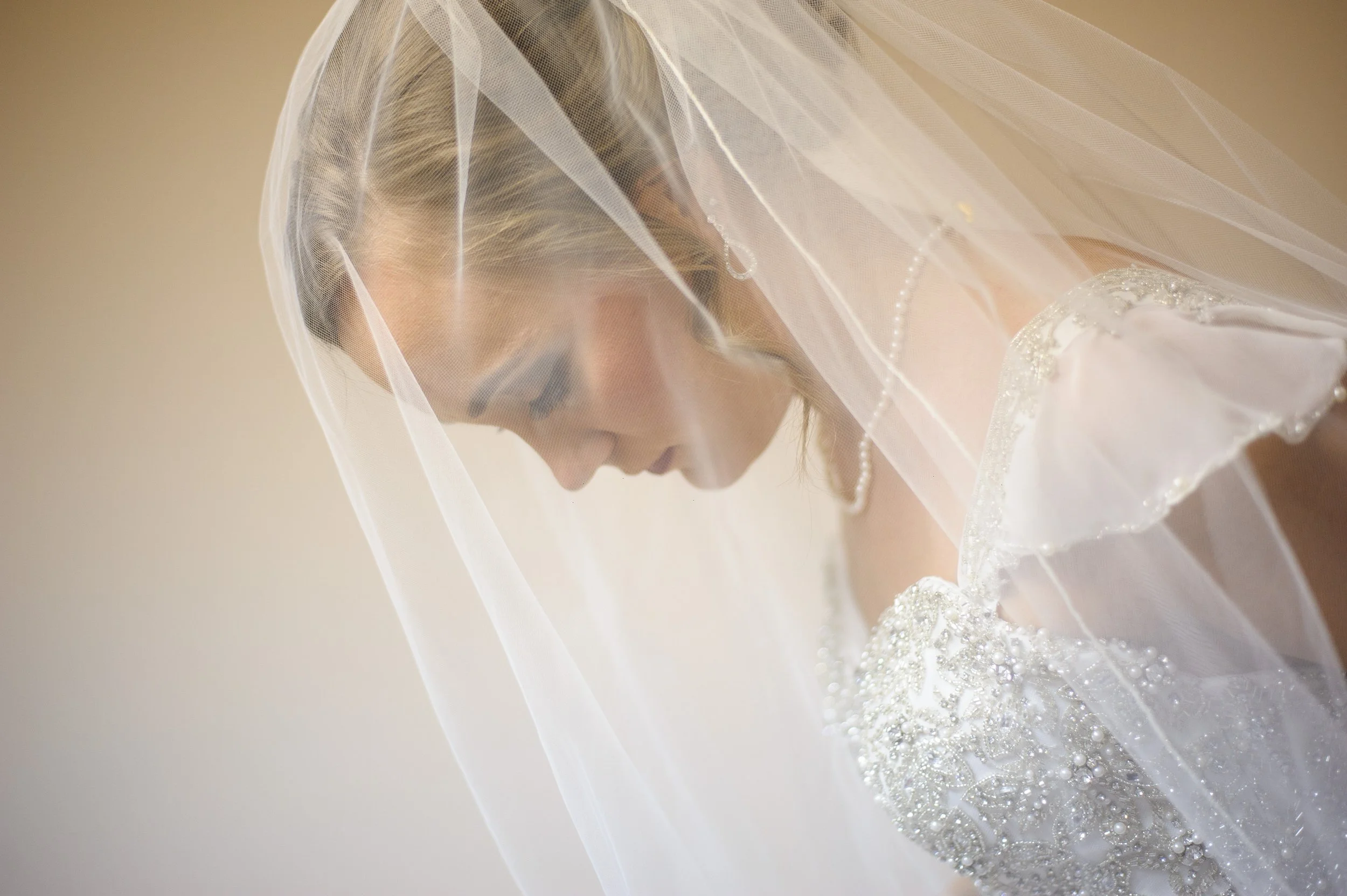 A bride in a wedding dress and veil looking down, with pearl jewelry and detailed beaded embroidery on her dress.