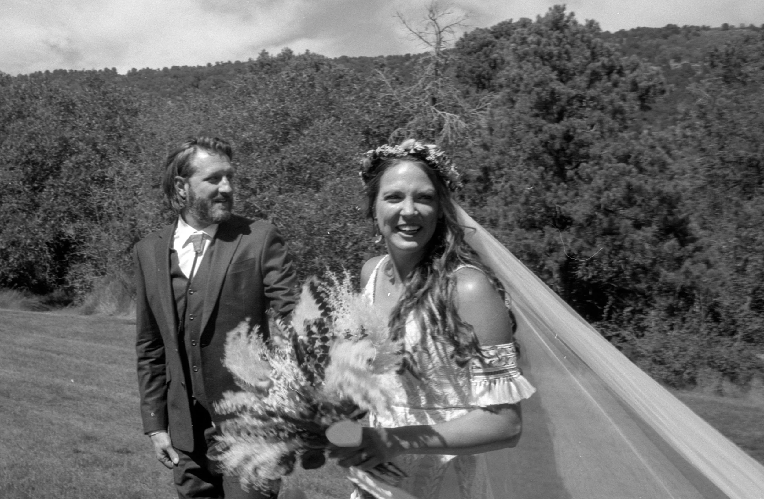 Black and white photo of a smiling bride holding a bouquet of flowers, wearing a floral crown and veil, with a groom in a suit standing beside her, outdoors with trees and hills in the background.