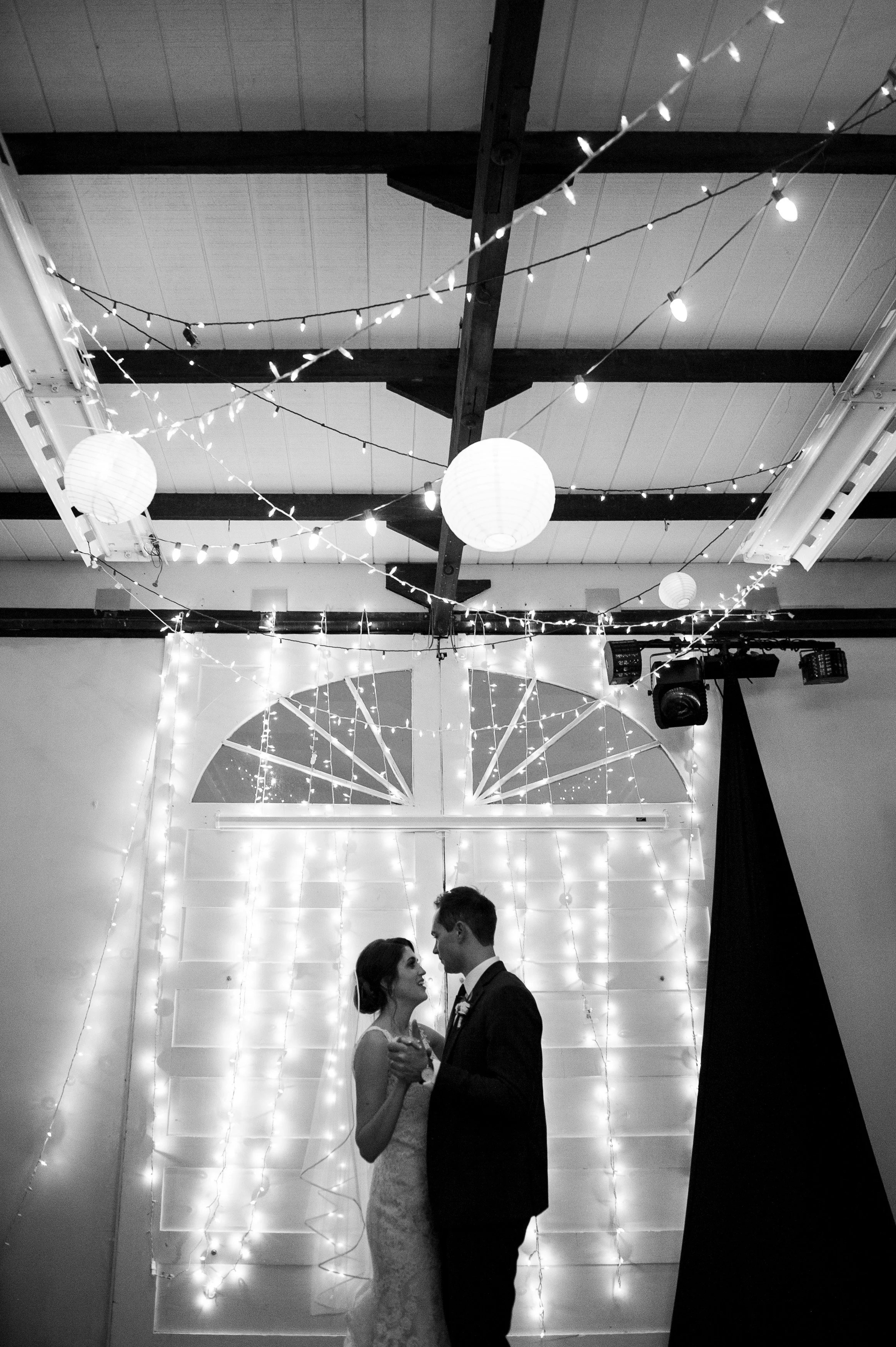 A black and white photo of a bride and groom dancing at their wedding reception with string lights and paper lanterns hanging from the ceiling.