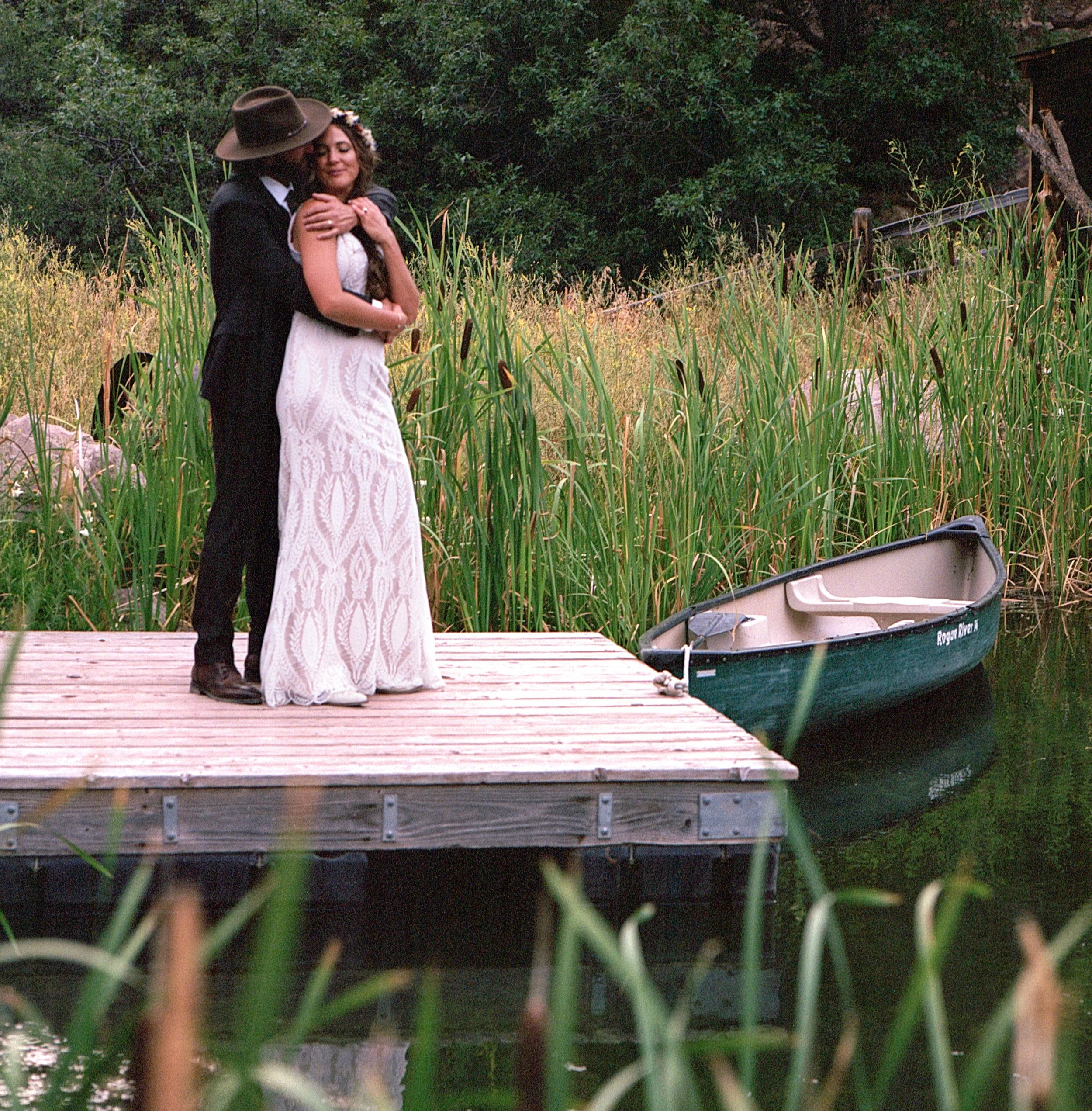 A couple embraces on a wooden dock near a boat, surrounded by tall grass and trees.