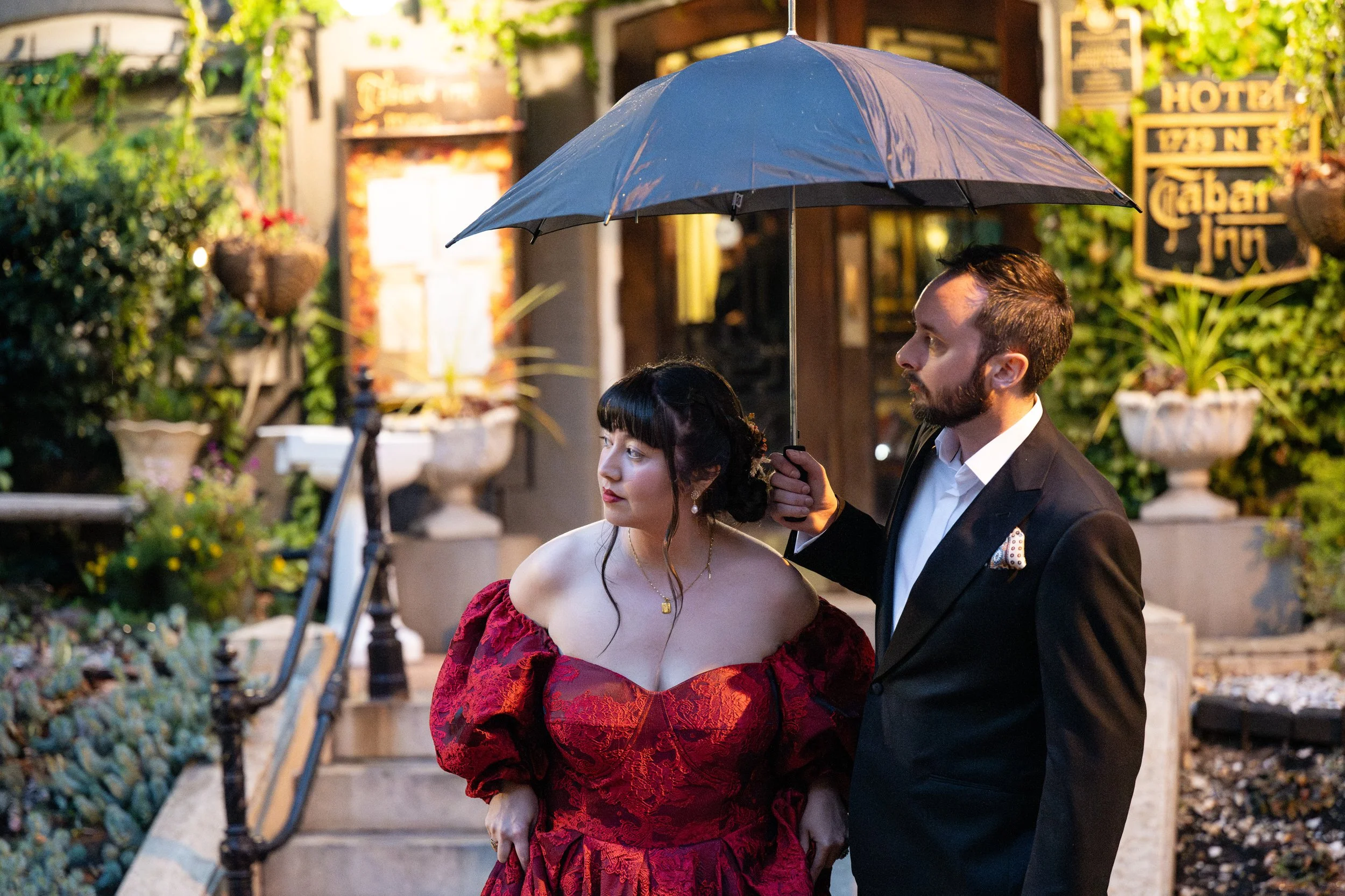 A man in a black suit holding a black umbrella over a woman in a red off-shoulder dress, standing outdoors near stairs and surrounded by greenery.