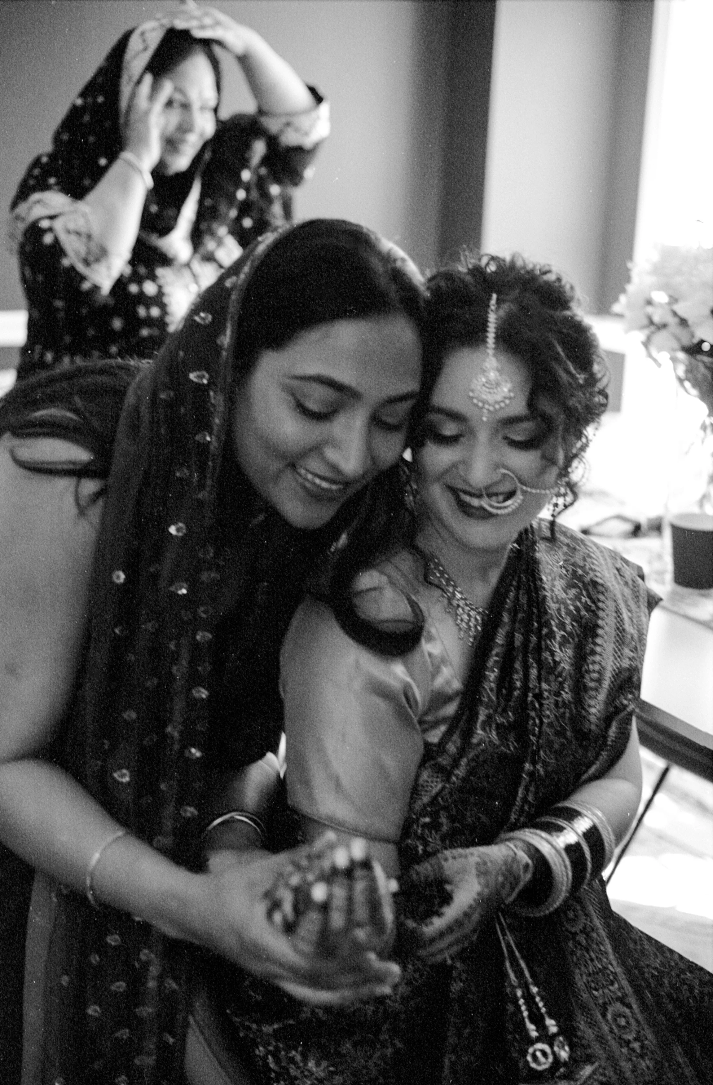 Two women dressed in traditional Indian attire smiling, with a third woman in the background also smiling and holding her head, inside a room with light coming through a window.