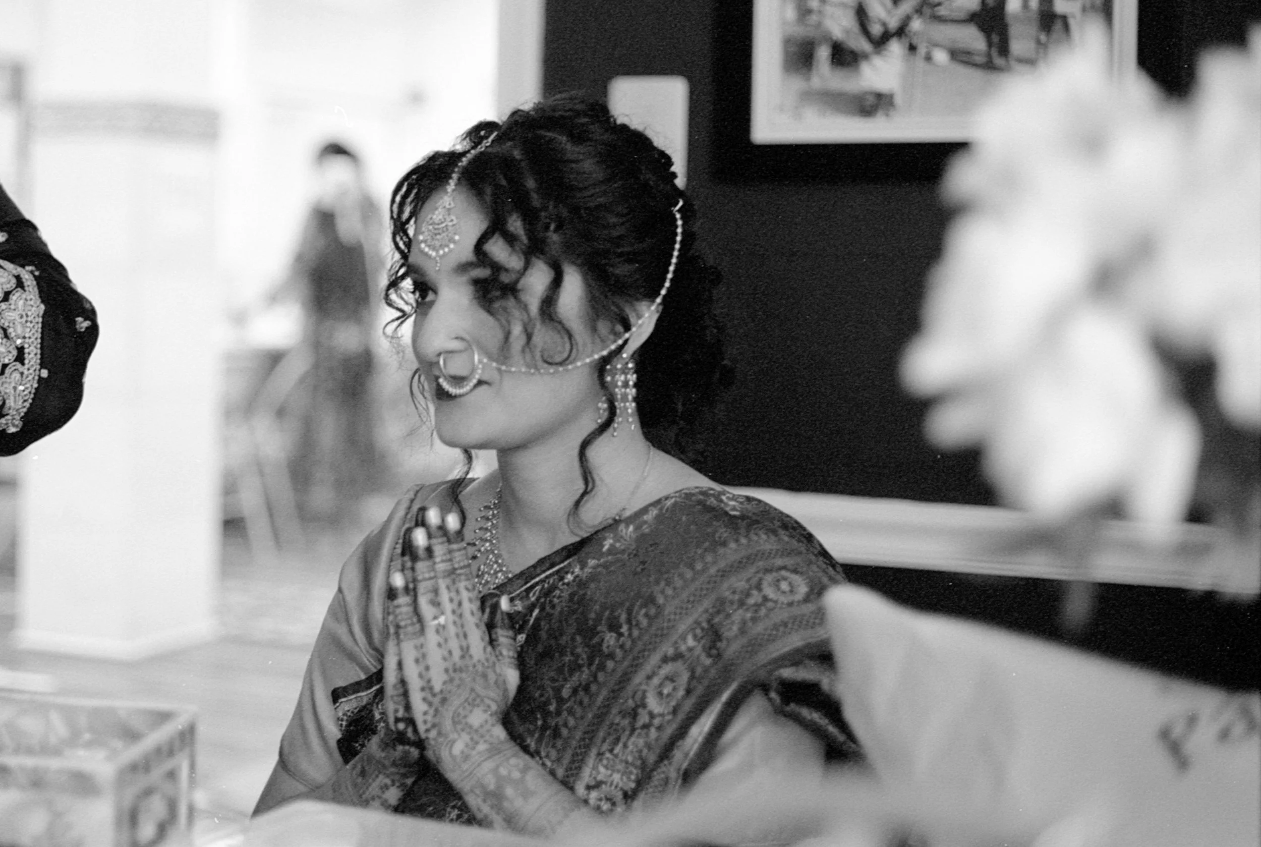A woman dressed in traditional Indian attire with intricate jewelry, including a nose ring, earrings, and a forehead ornament. She has decorated hands with henna and her eyes are closed, appearing to be in prayer or meditation.