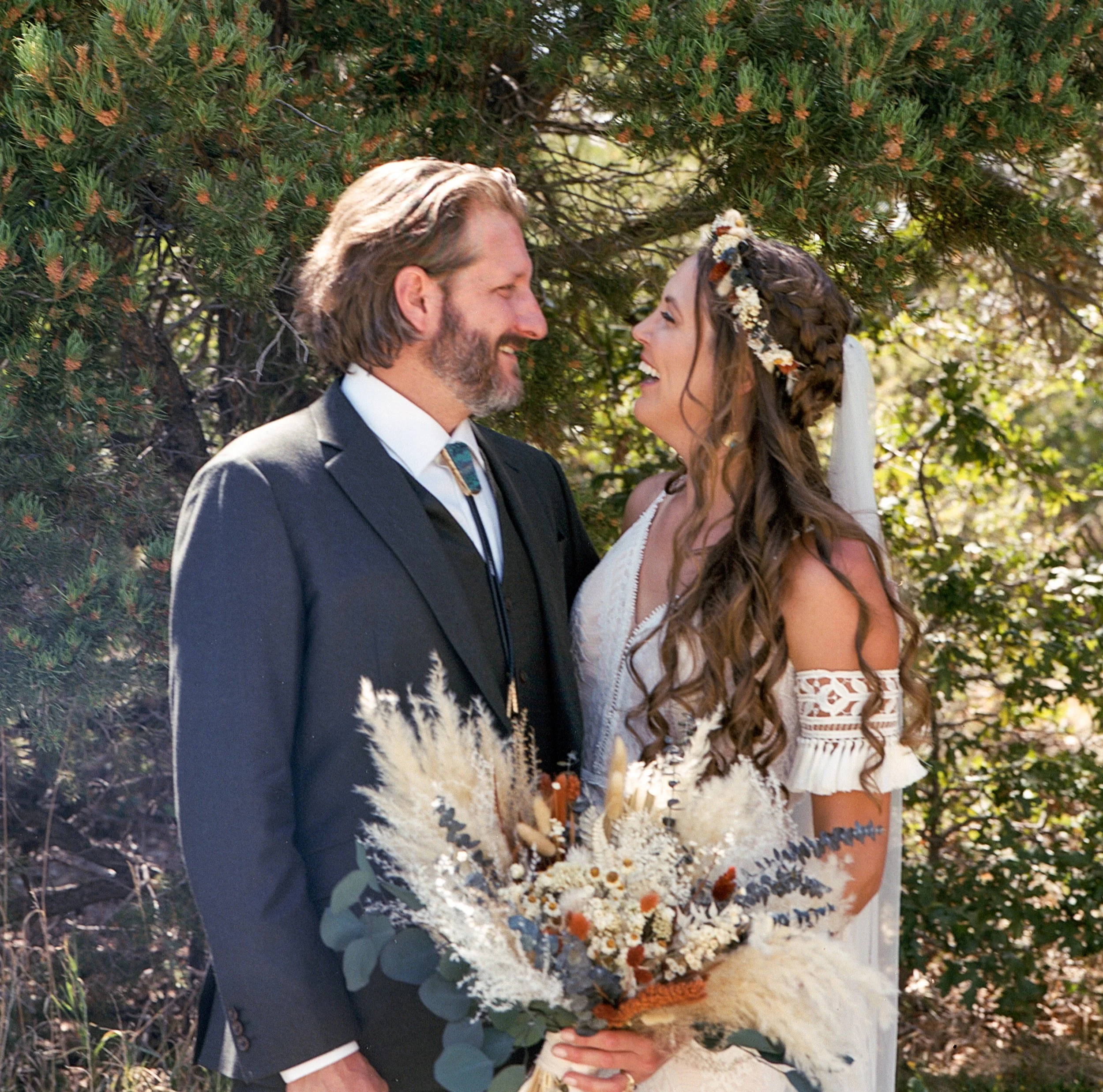 A bride and groom standing outdoors, smiling at each other, with trees in the background. The bride has long, curly hair, a floral headband, and is holding a bouquet.