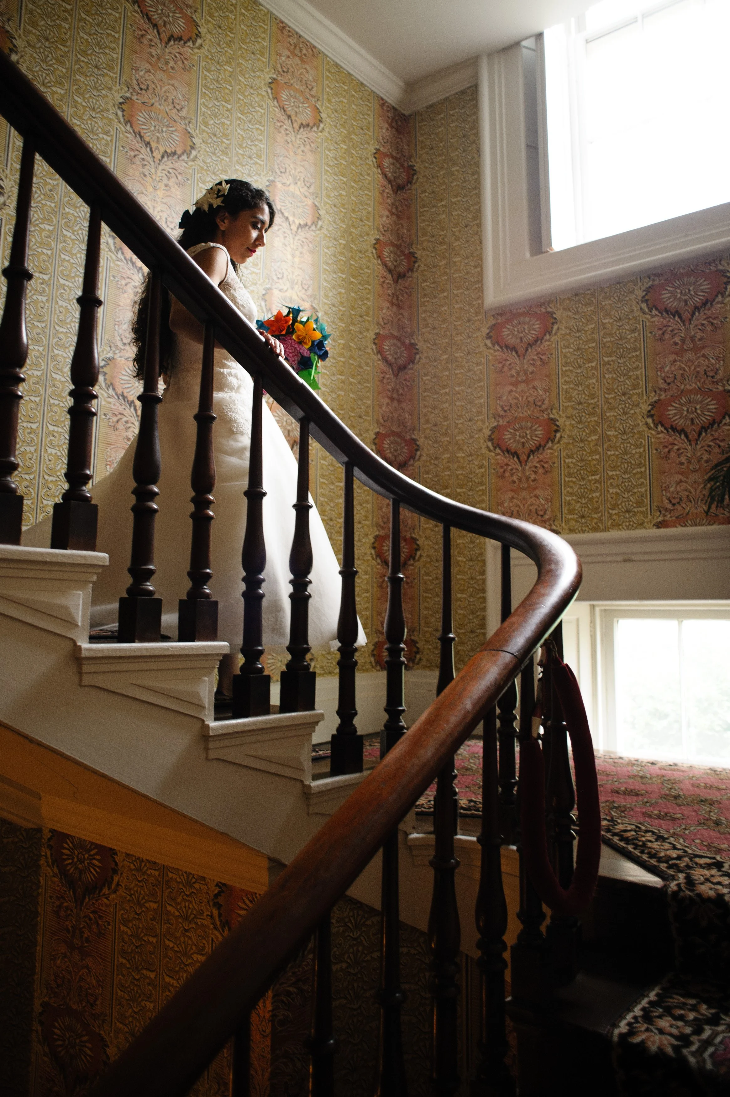 A woman in a wedding dress holding a bouquet of colorful flowers on a staircase with ornate wallpaper and wooden banister.