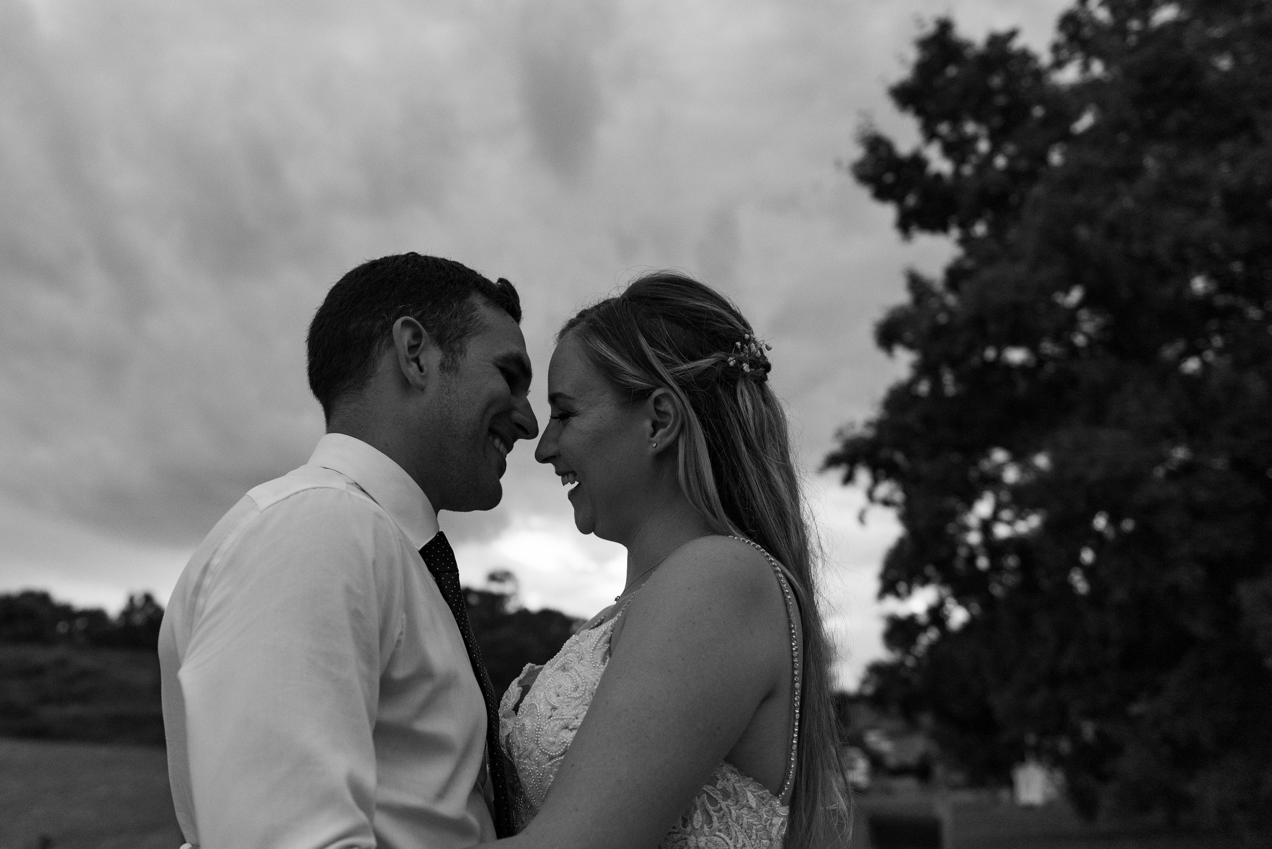 A black-and-white photo of a couple embracing and touching foreheads outdoors, smiling happily, dressed in wedding attire.