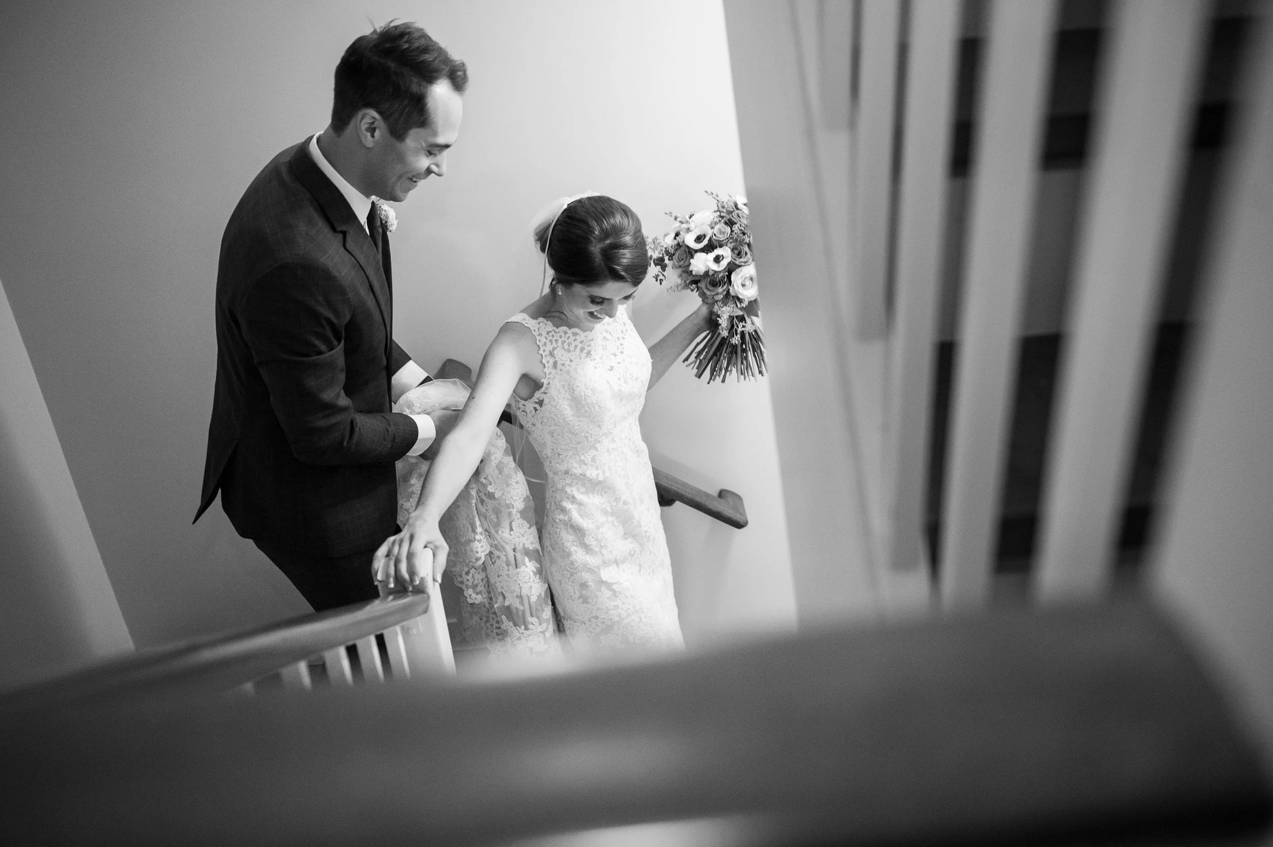 A black-and-white photo of a bride and groom descending a staircase together, with the bride holding a bouquet and smiling, and the groom assisting her.