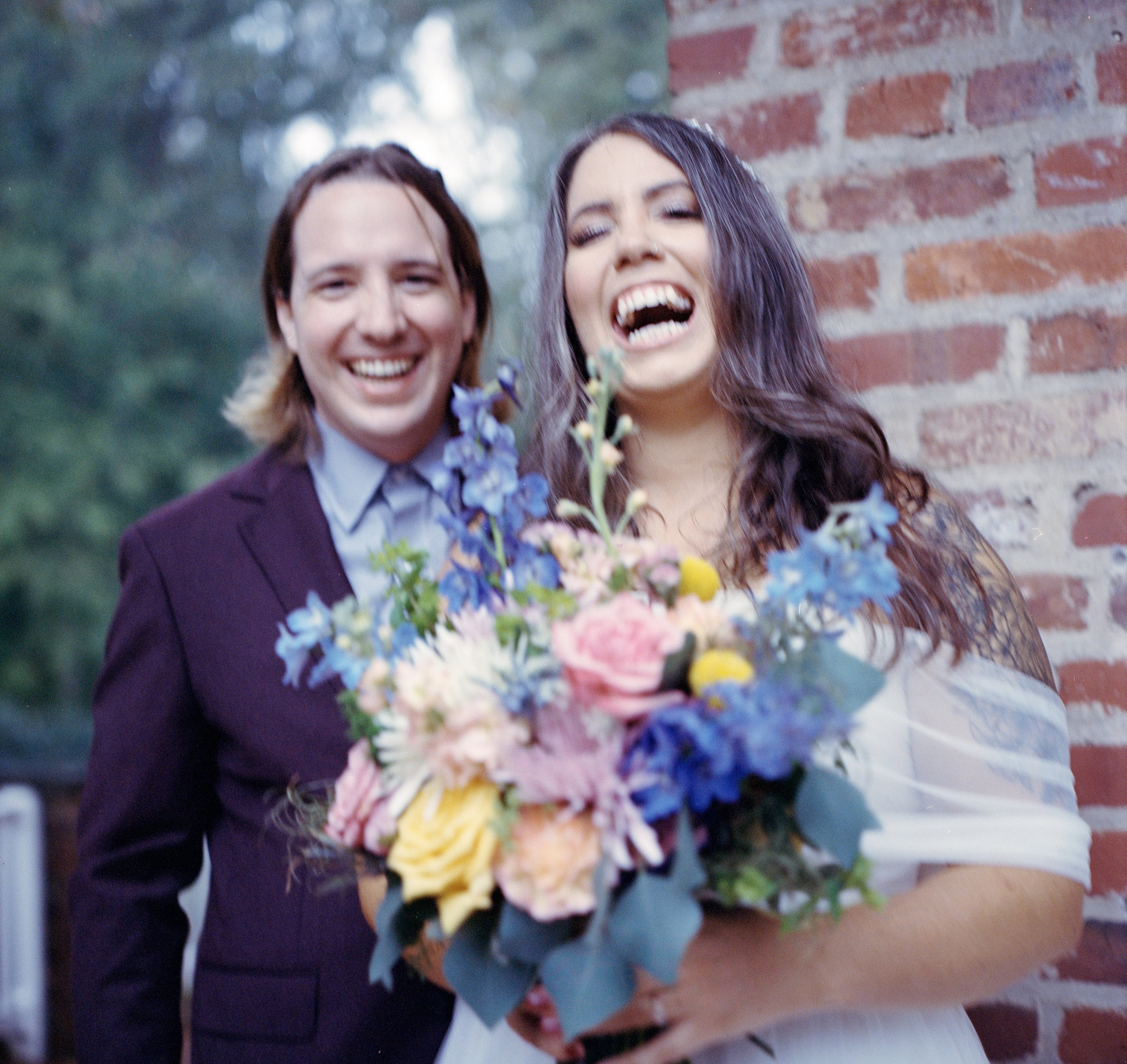 A couple smiling, with the woman holding a bouquet of colorful flowers, standing outside near a brick wall.