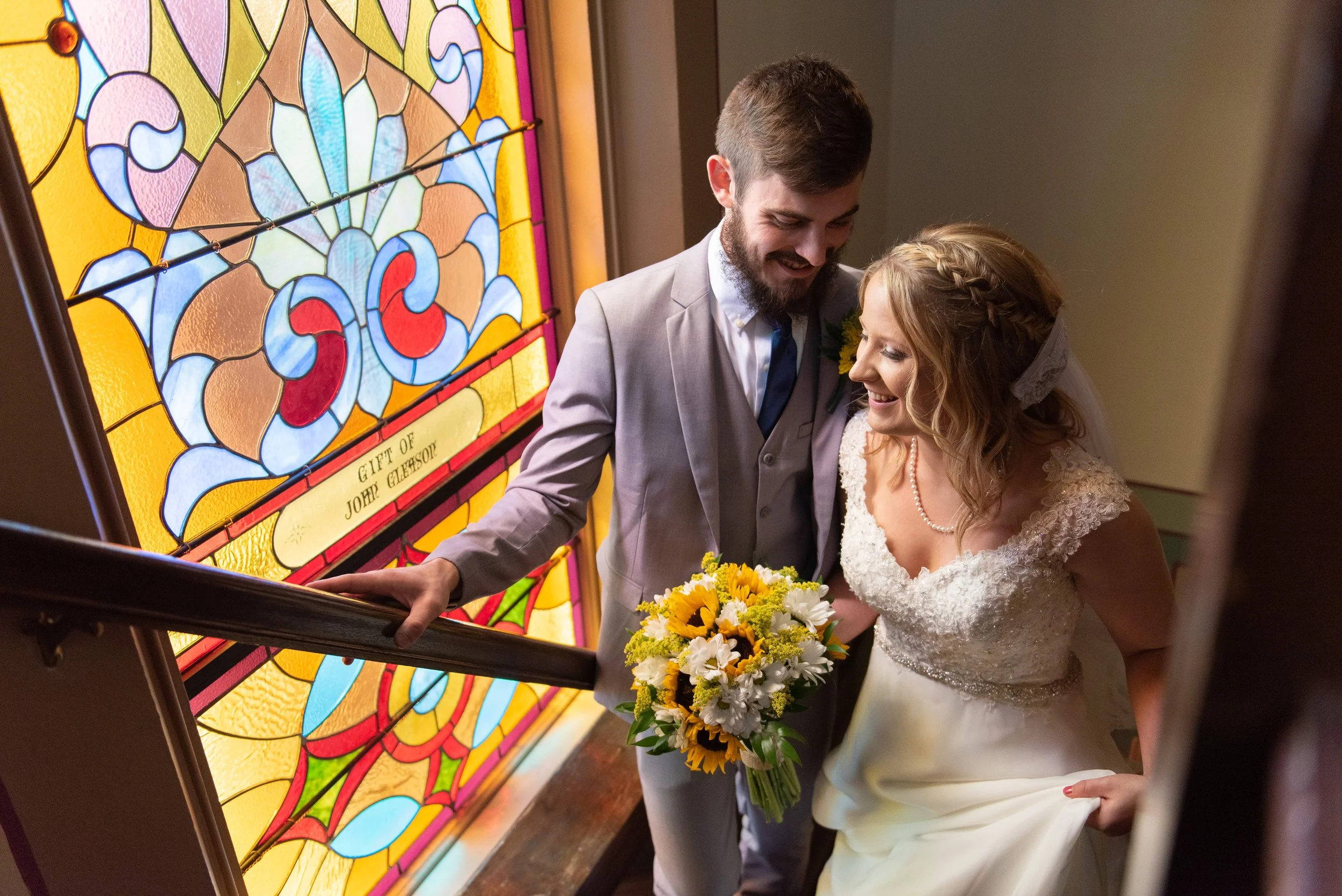A newlywed couple, a man in a light gray suit and a woman in a wedding dress, sharing a joyful moment as they descend a staircase. The bride is holding a bouquet of sunflowers and white flowers, and a colorful stained glass window is visible in the b