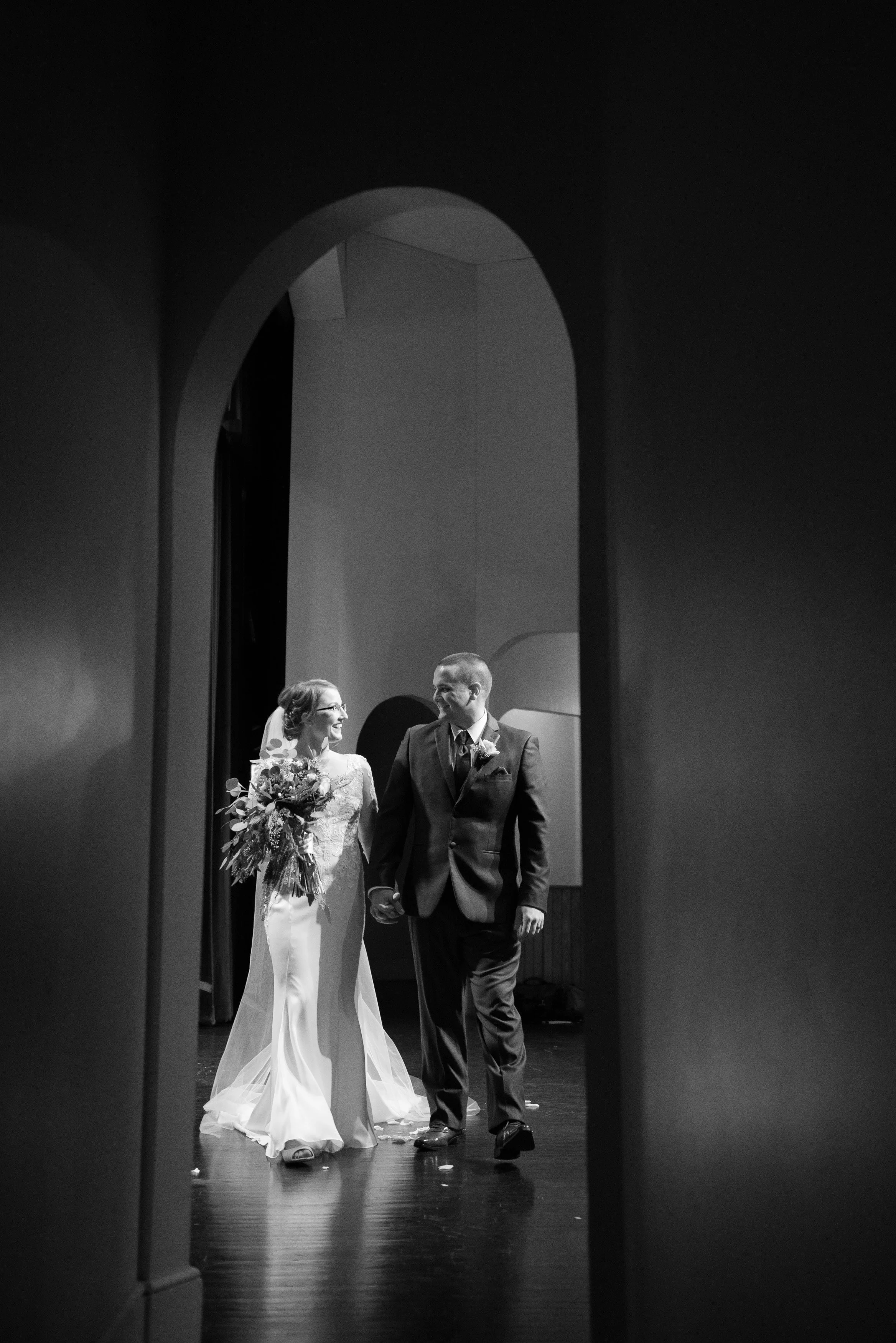 Black and white photo of a bride and groom holding hands and walking together, seen through an archway, smiling at each other.