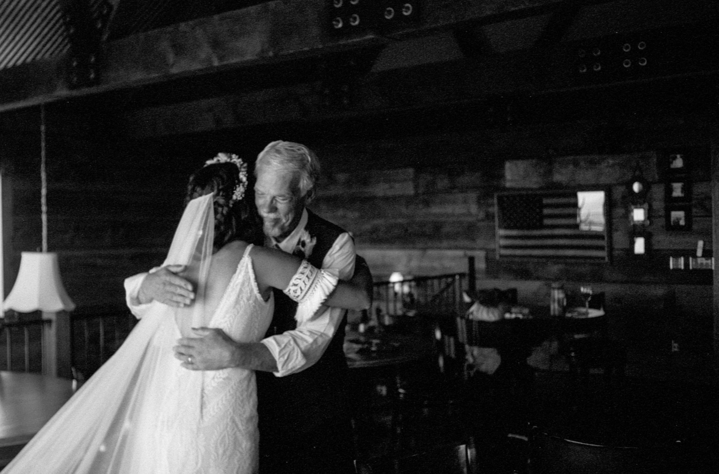 An elderly man and a woman in a wedding dress sharing a warm hug inside a rustic wooden room, with American flags and framed pictures on the wall in the background.