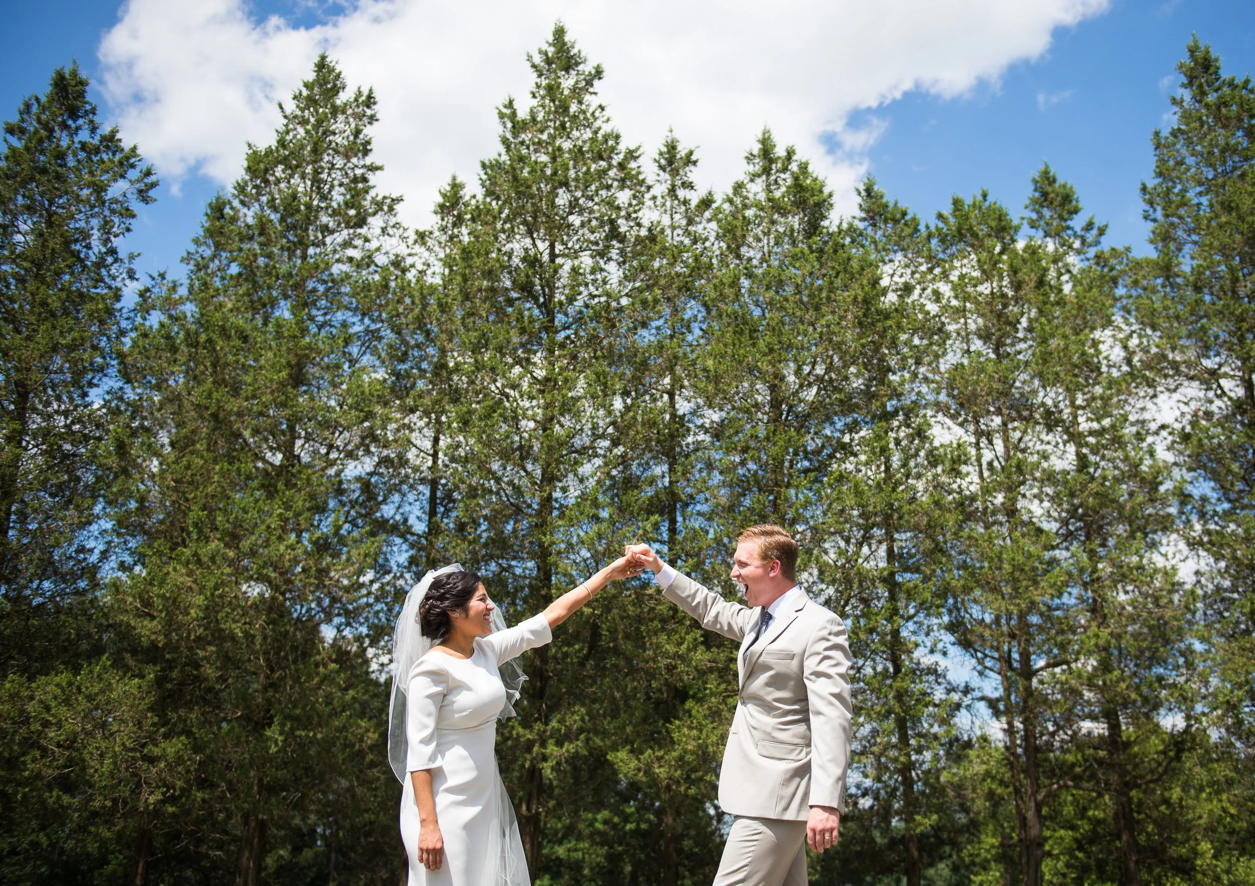 A bride and groom smiling and dancing outdoors under a blue sky with clouds, surrounded by green trees.