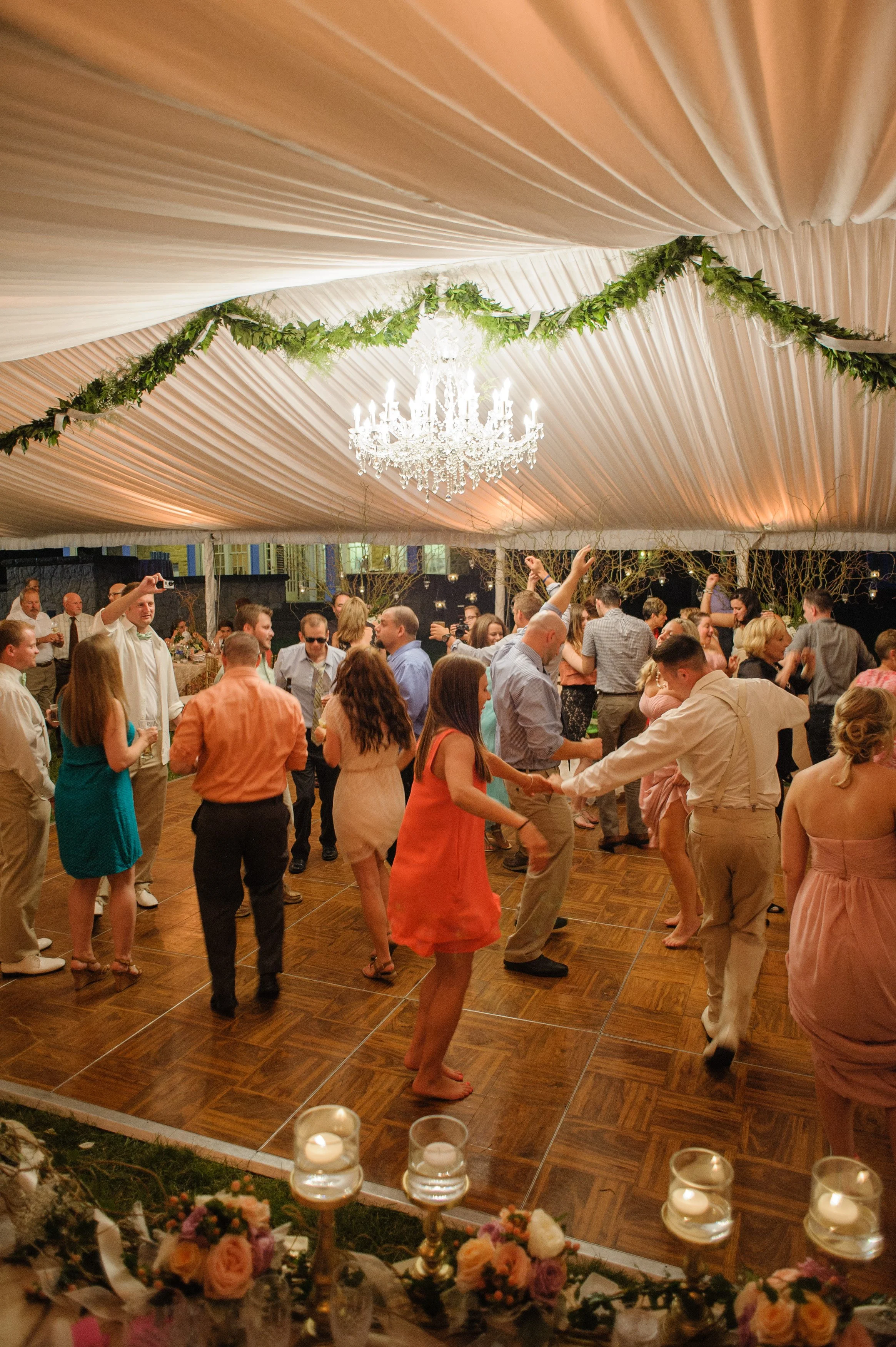People dancing and celebrating at a wedding reception under a decorated tent ceiling with a chandelier, greenery, and candles on a table in the foreground.