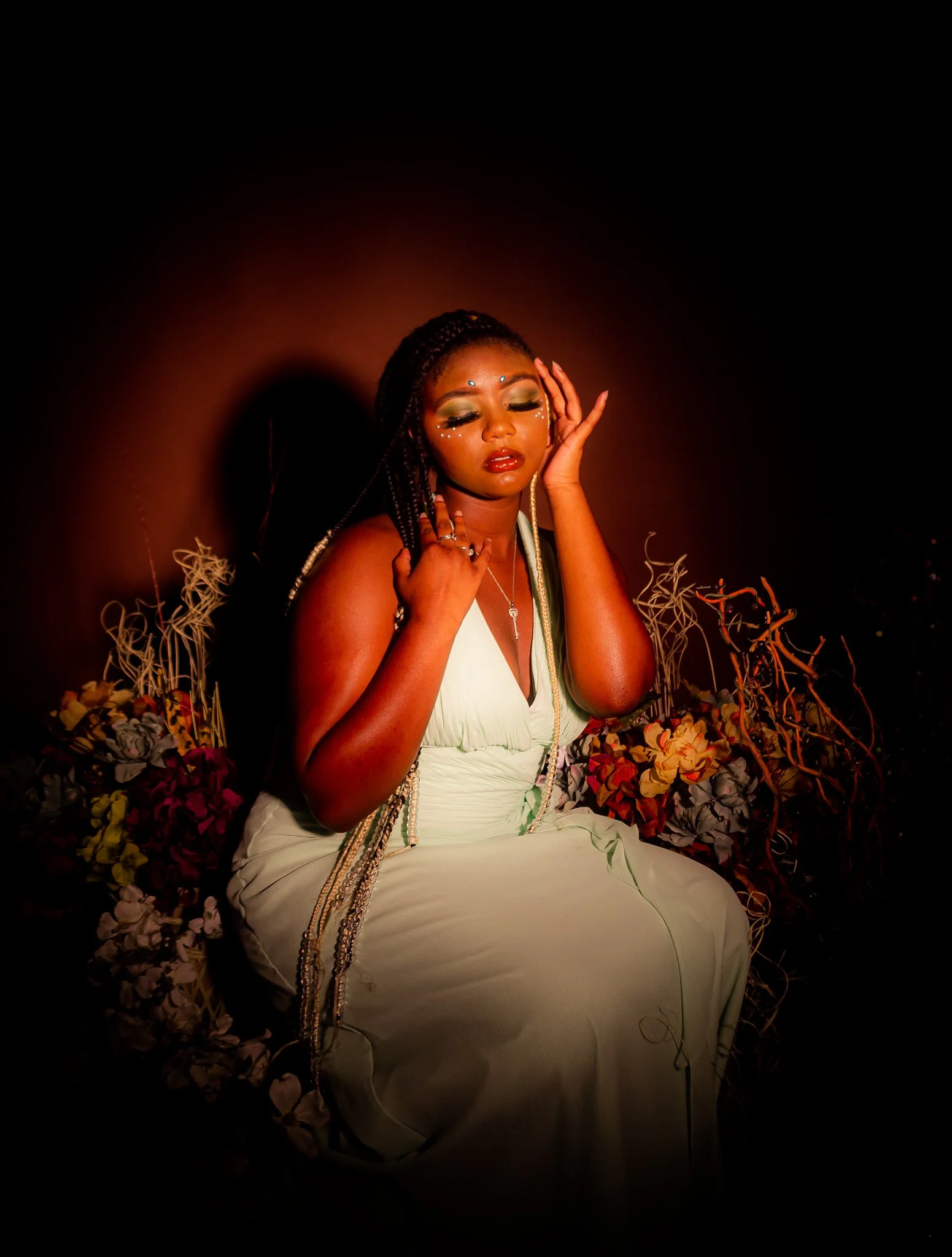 A woman with braided hair and makeup, dressed in a white gown, sitting among flowers with closed eyes and her hand touching her face.