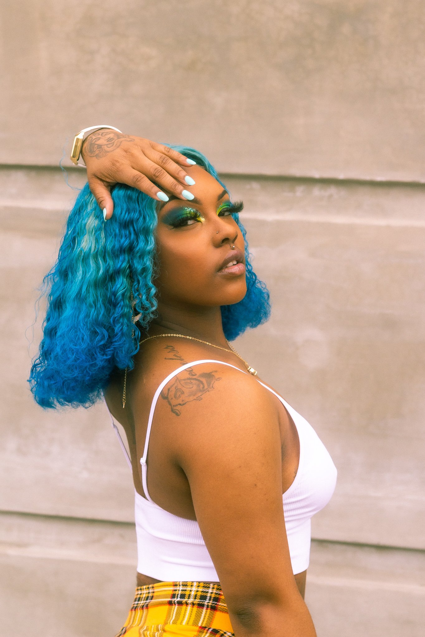 A woman with vibrant blue curly hair, wearing a white tank top and plaid yellow skirt, posing with her hand on her forehead, showcasing makeup, tattoos, and jewelry.