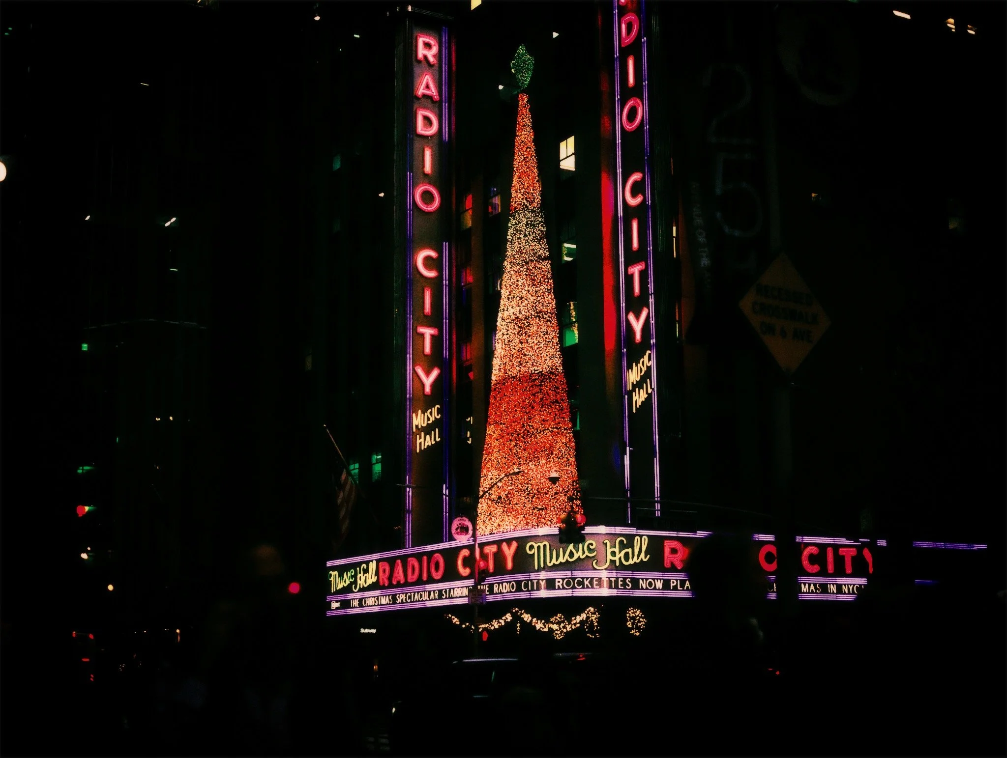 Neon signs on a building reading 'Radio City' and 'Music Hall' with a large illuminated Christmas tree between them at night in New York City.