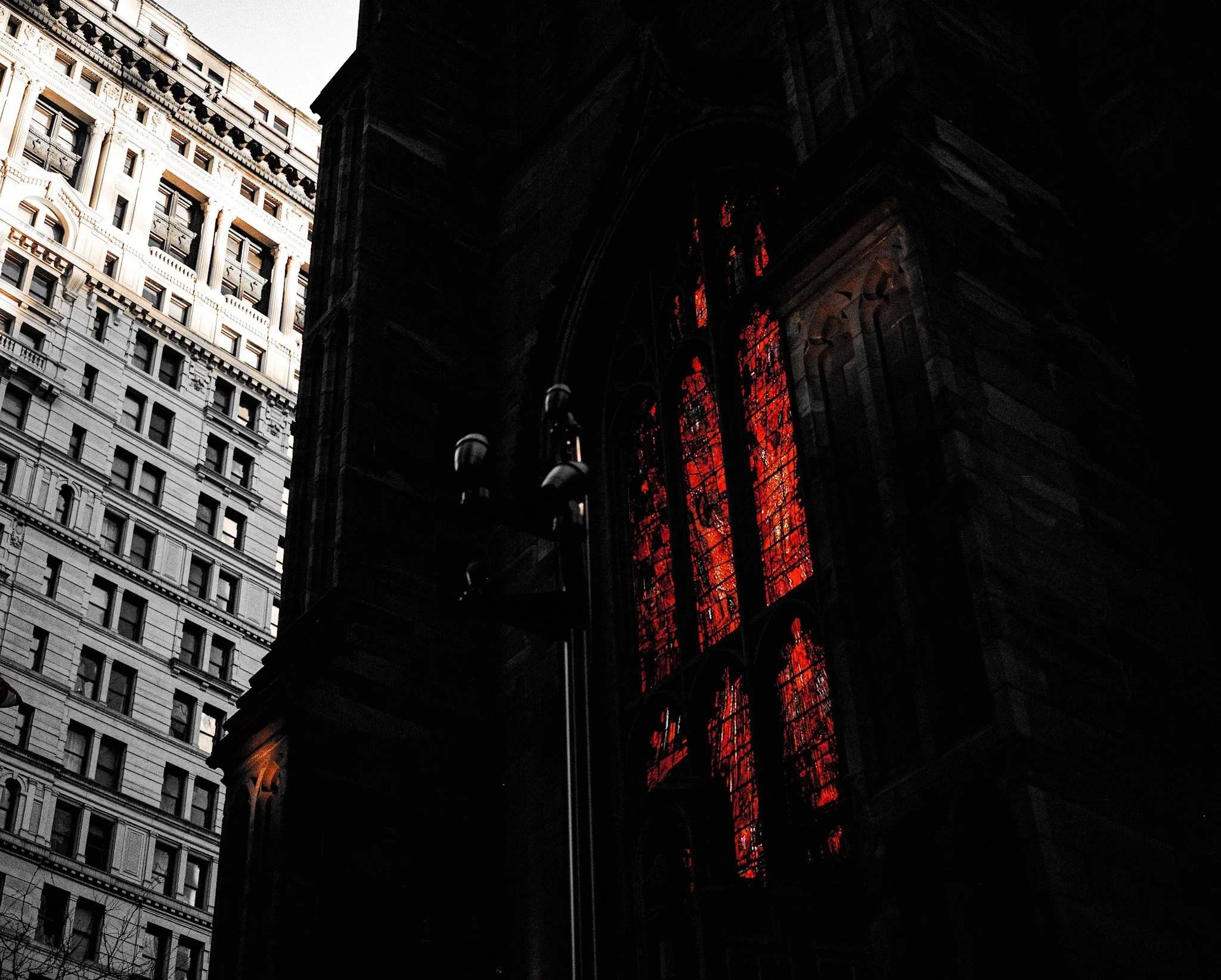 A dark church interior with red stained glass windows illuminated from inside. Modern high-rise buildings are visible outside through the window, contrasting the dark interior.