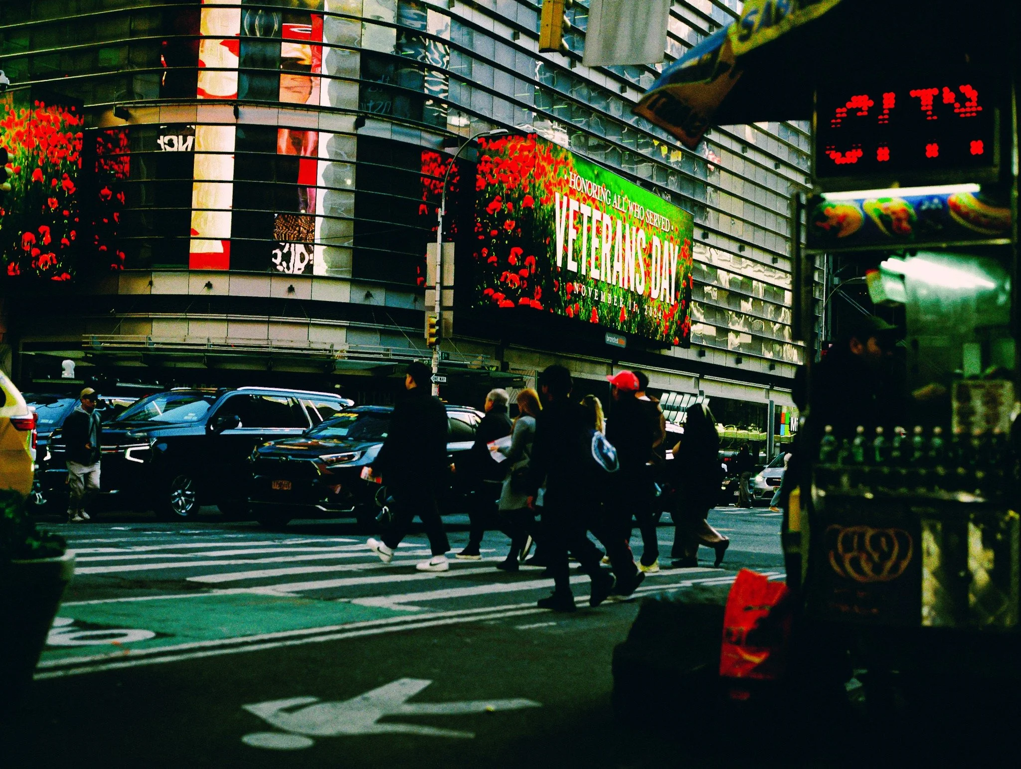 People walking across a crosswalk in Times Square, New York City, with a large digital screen displaying a Veterans Day advertisement and surrounding buildings and vehicles.