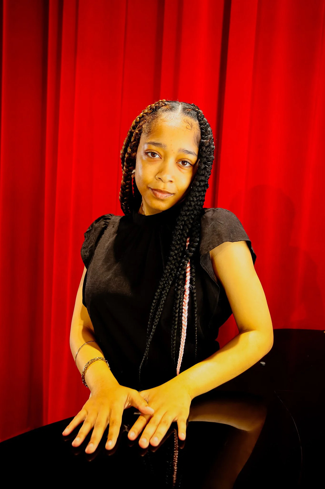 A young woman with braided hair wearing a black top, standing in front of a red curtain, with hands resting on a reflective black surface.