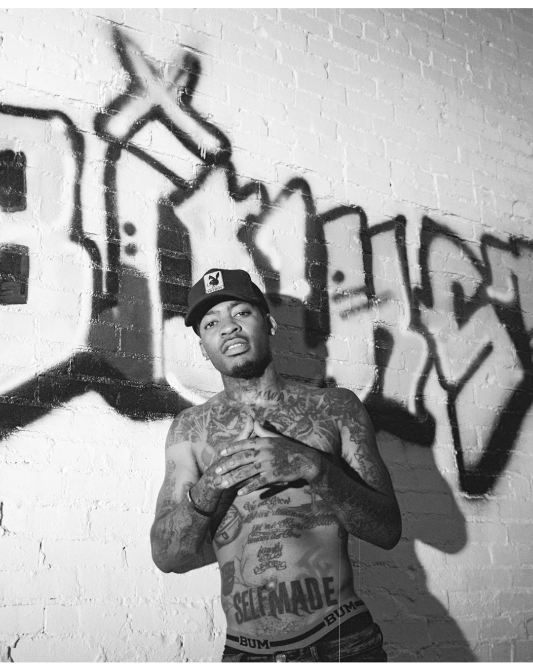 A shirtless man covered in tattoos standing against a brick wall with graffiti art in the background. The graffiti includes a large hand making a gesture and the word "BOY." The man is wearing a baseball cap and underwear, with the word "SELFMADE" vi