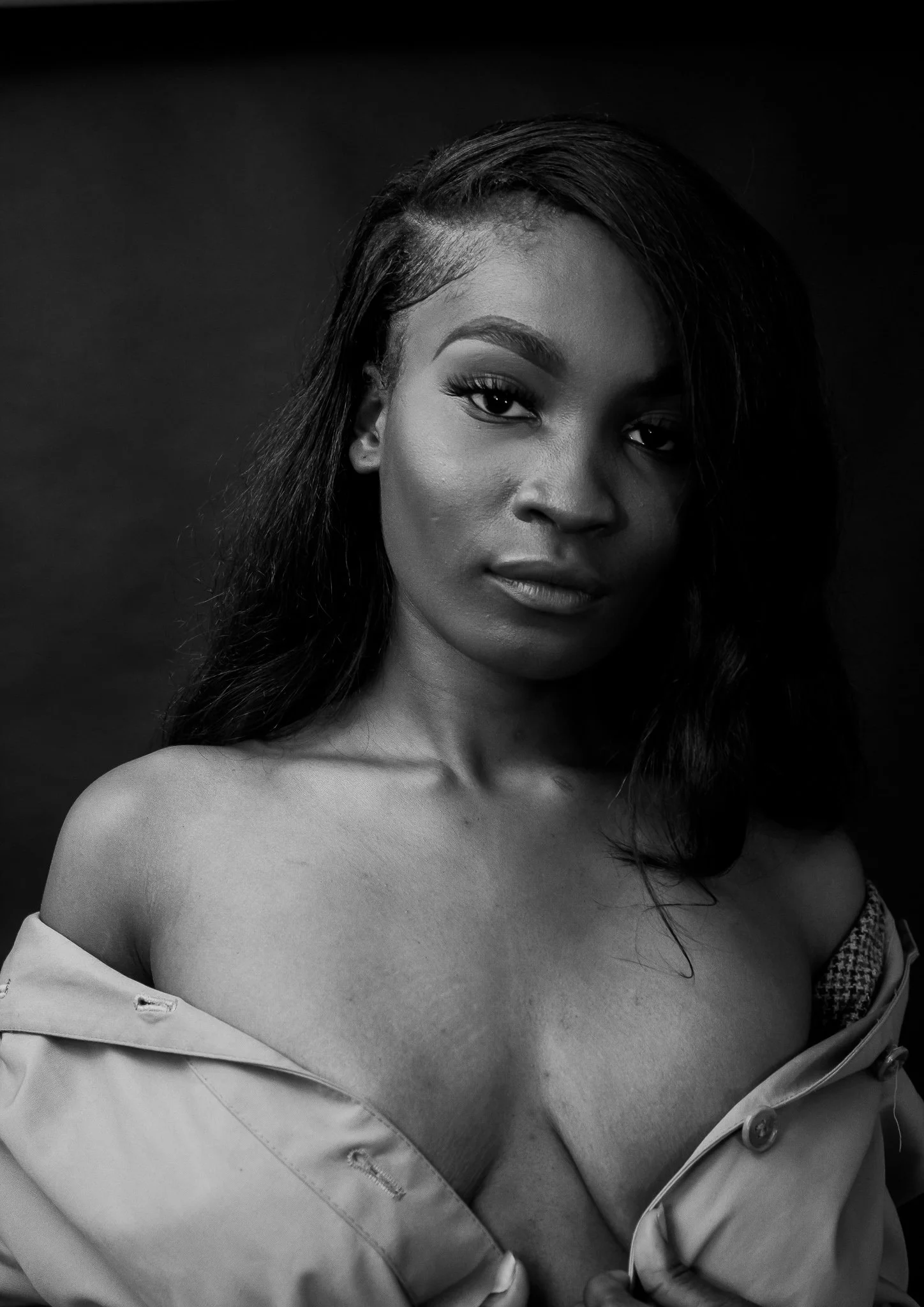 Black and white portrait of a young woman with long hair, partially covered by an off-the-shoulder coat, looking directly at the camera.