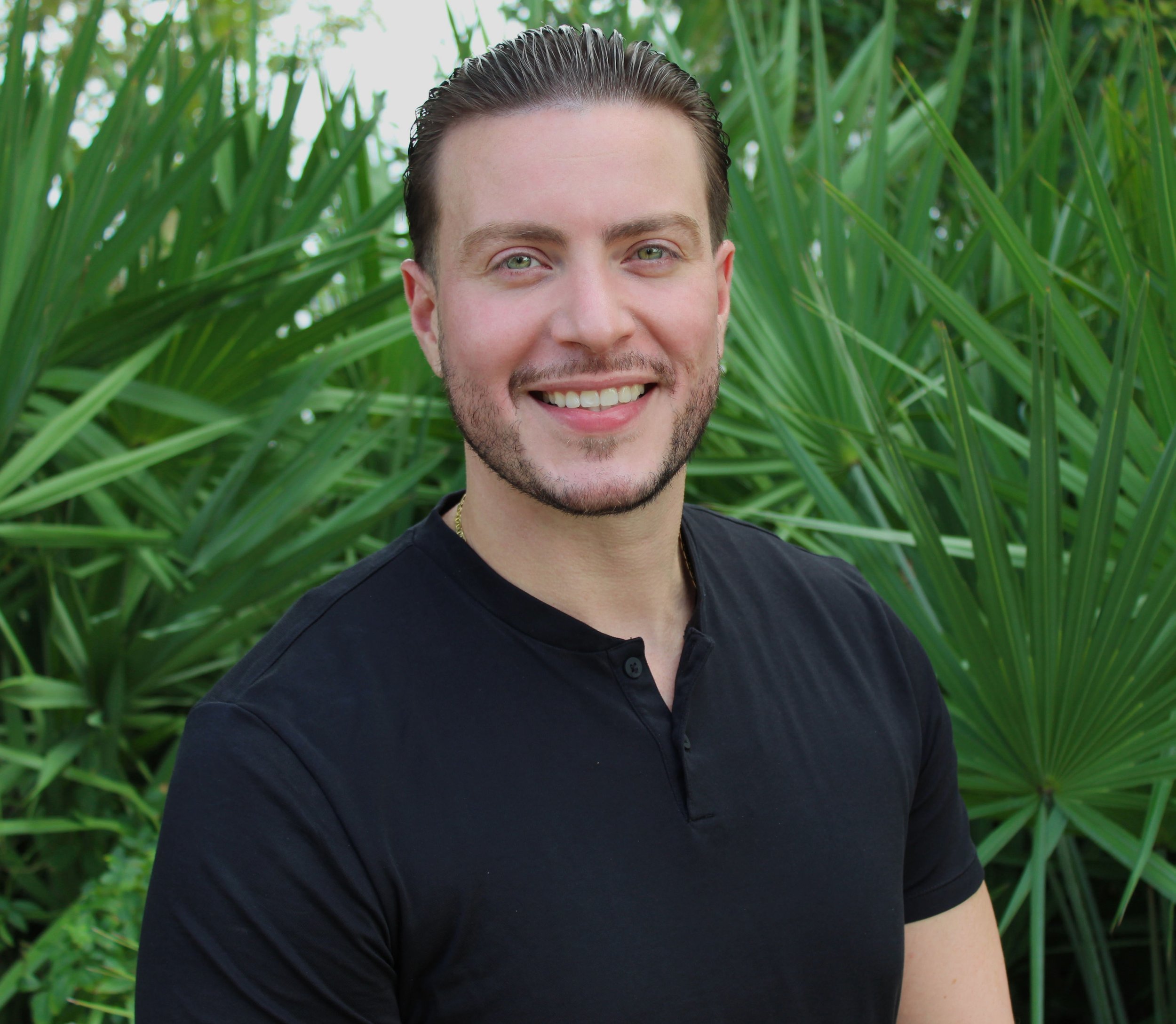 A smiling man with light skin, short brown hair, and a beard, wearing a black shirt, standing in front of green plants.