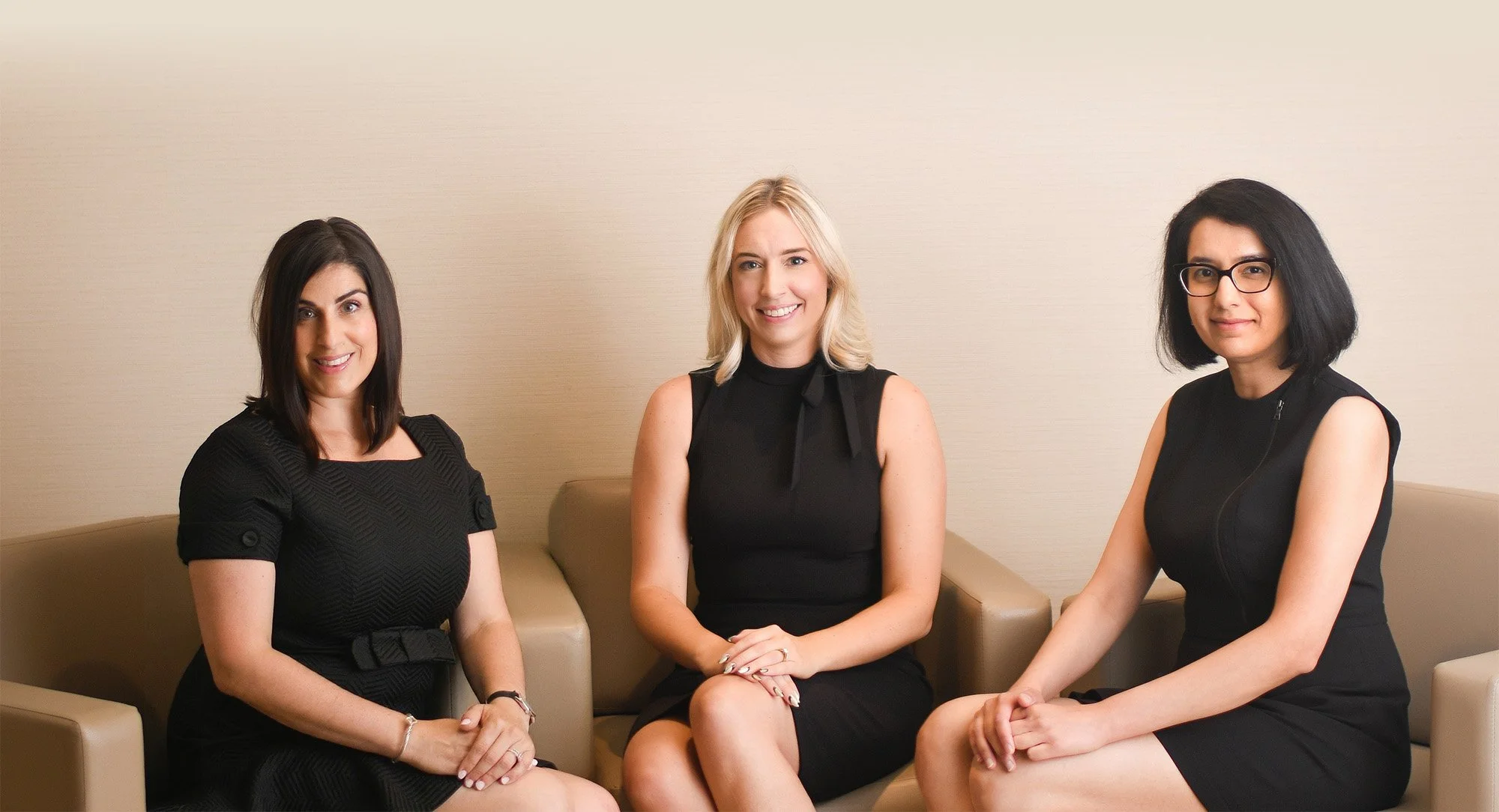 Three women: Laura Natalizio, Heather Cassells, and Nida Hussein, dressed in black professional attire sitting on a beige sofa against a plain wall, smiling at the camera.