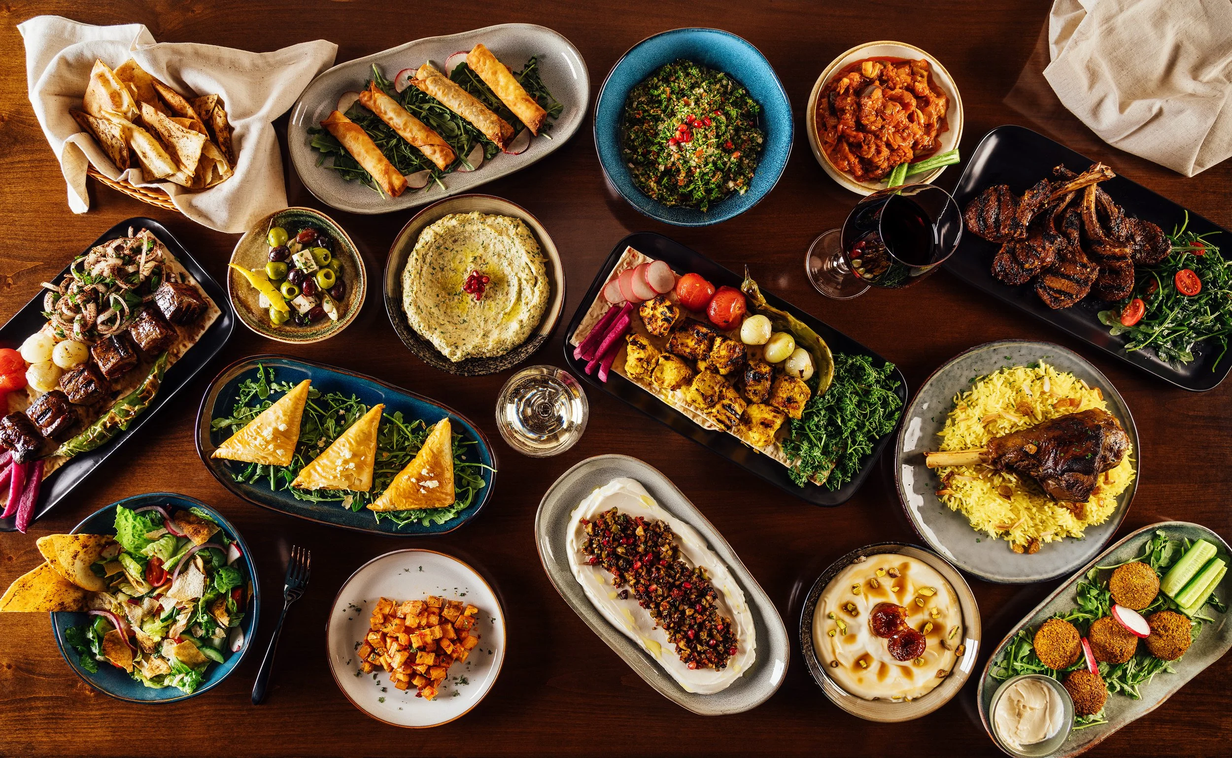 A large spread of various Middle Eastern dishes on a wooden table, including salads, dips, grilled meats, bread, and rice.