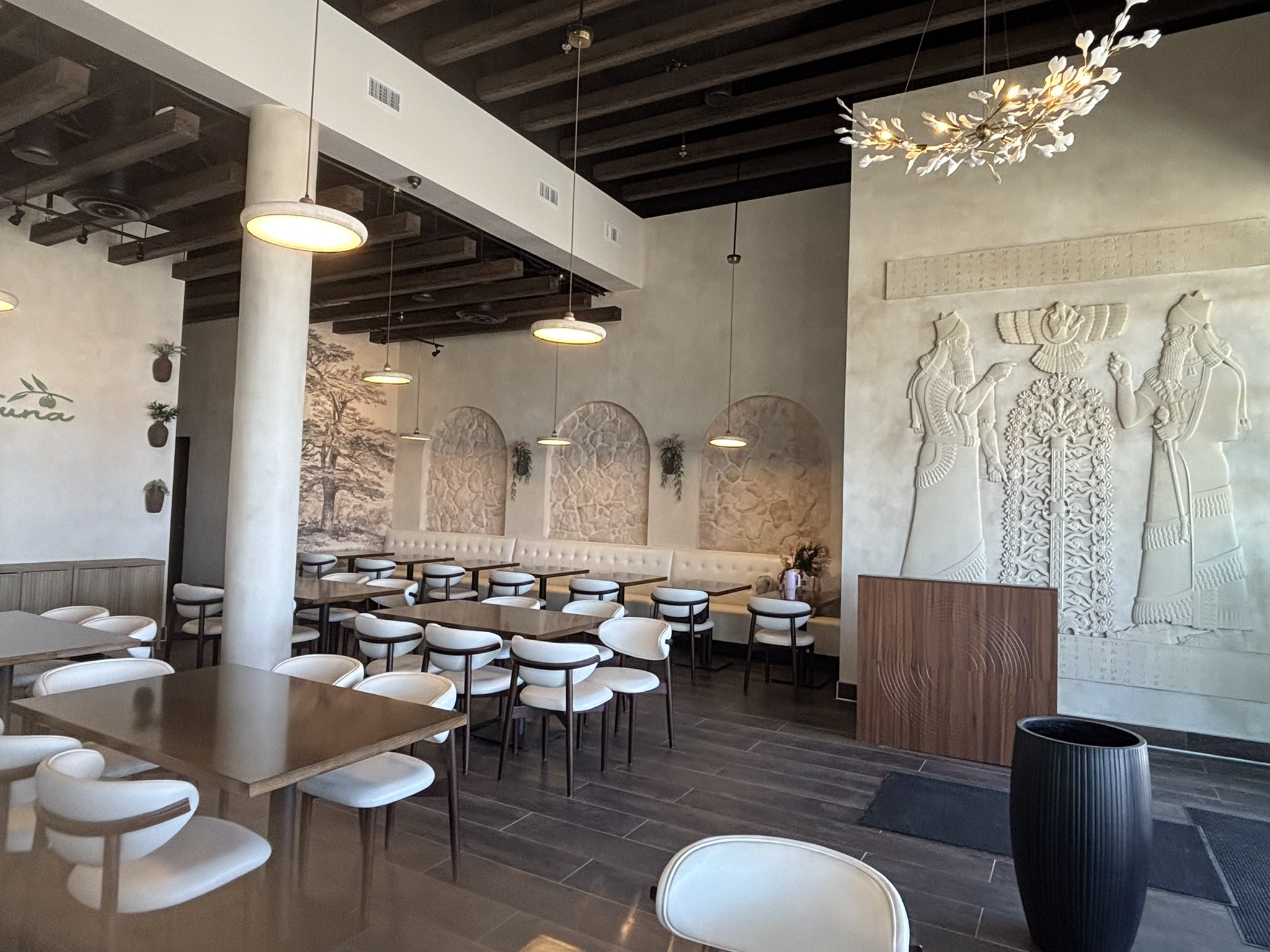 Interior of Zeitouna restaurant with wooden tables, white chairs, decorative wall art, hanging lights, and a textured wall with sculptures.