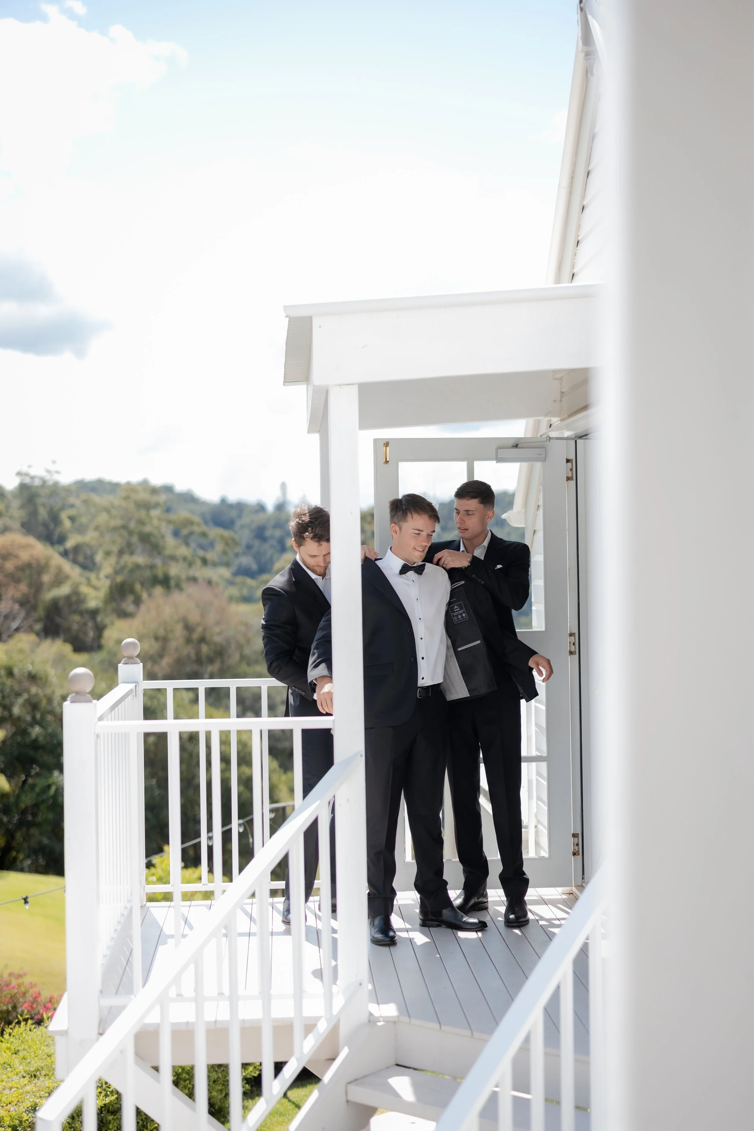 Three men in tuxedos preparing for a formal event on a balcony with a scenic view of trees in the background.
