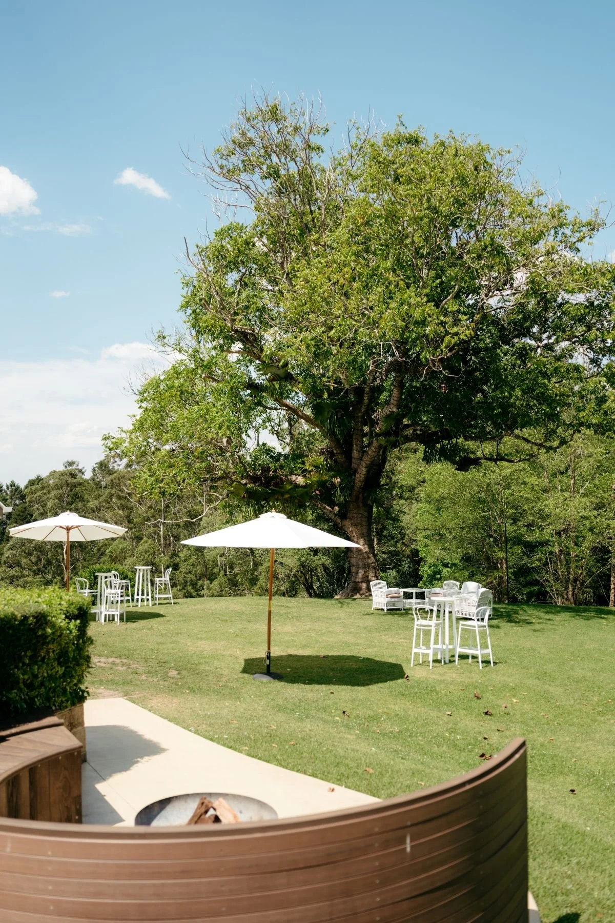 Outdoor garden party with white tables and chairs, shaded by large white umbrellas, lush green lawn, big tree, and a clear blue sky, and a curved fireplace.