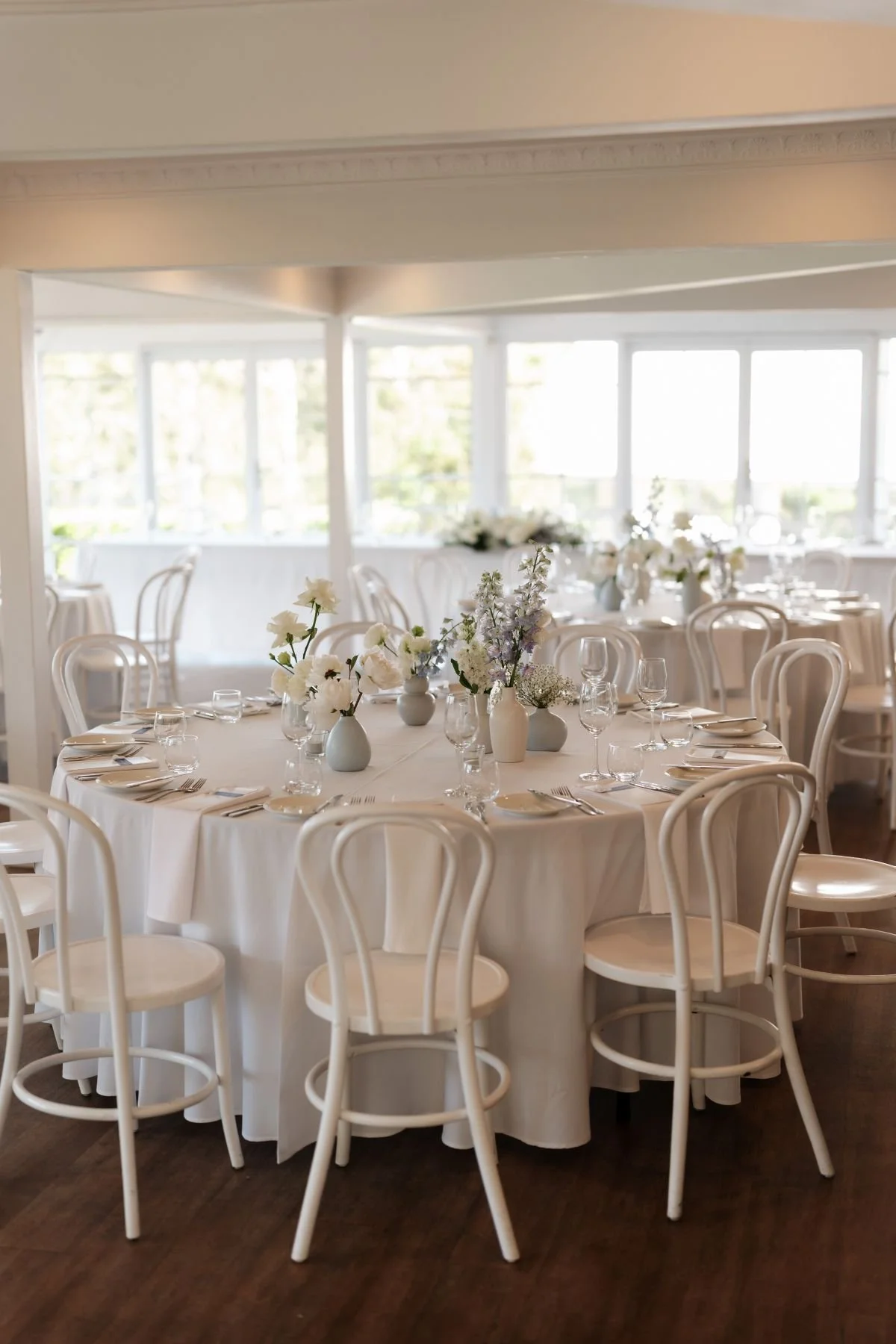 Elegant round table with white tablecloth, glassware, plates, silverware, and floral centerpieces in white vases, set in a bright, airy room with floor to ceiling windows.