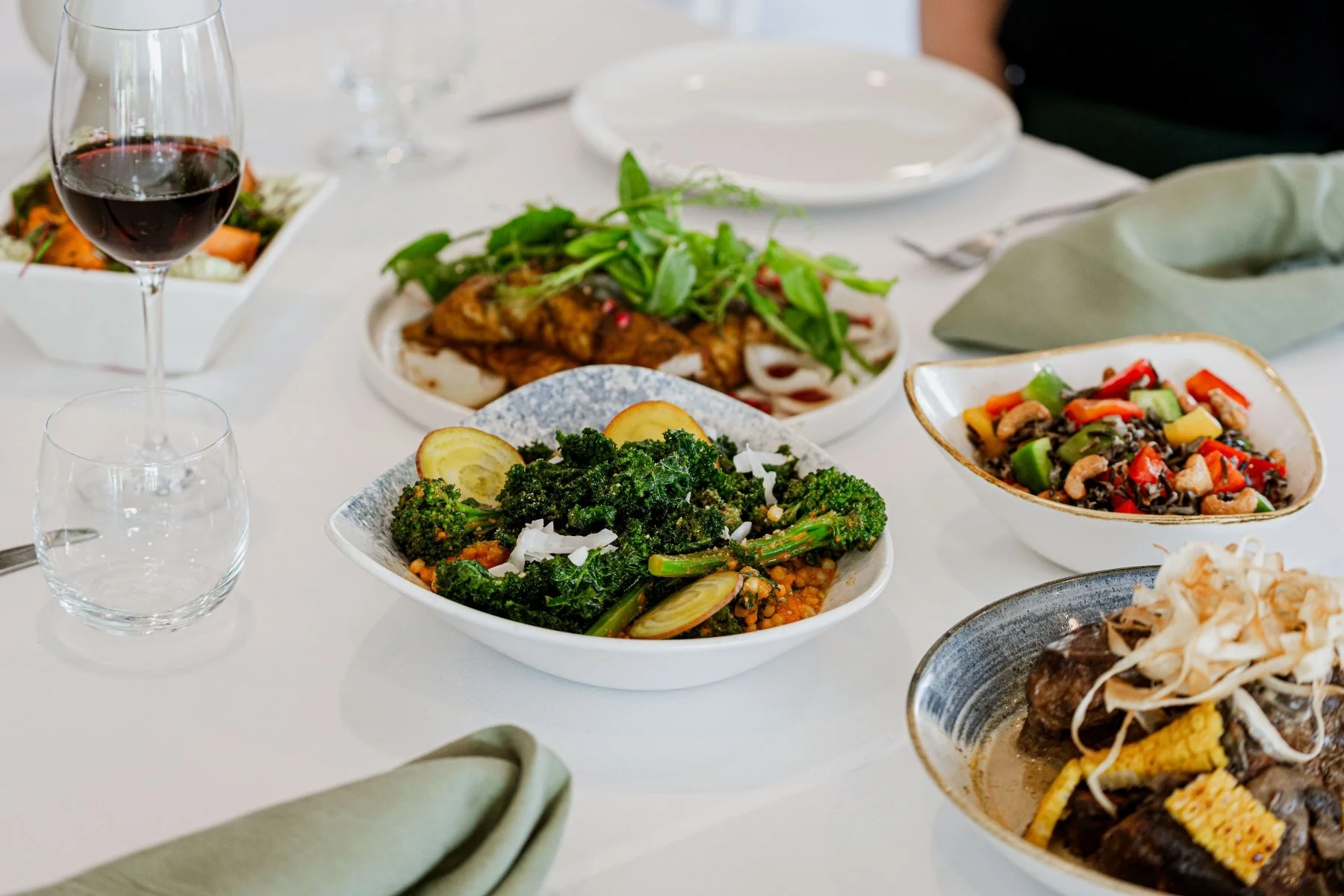 A table with various dishes including a glass of red wine, bowls of vegetables, salads, and a main course, set with plates, utensils, and cloth napkins.
