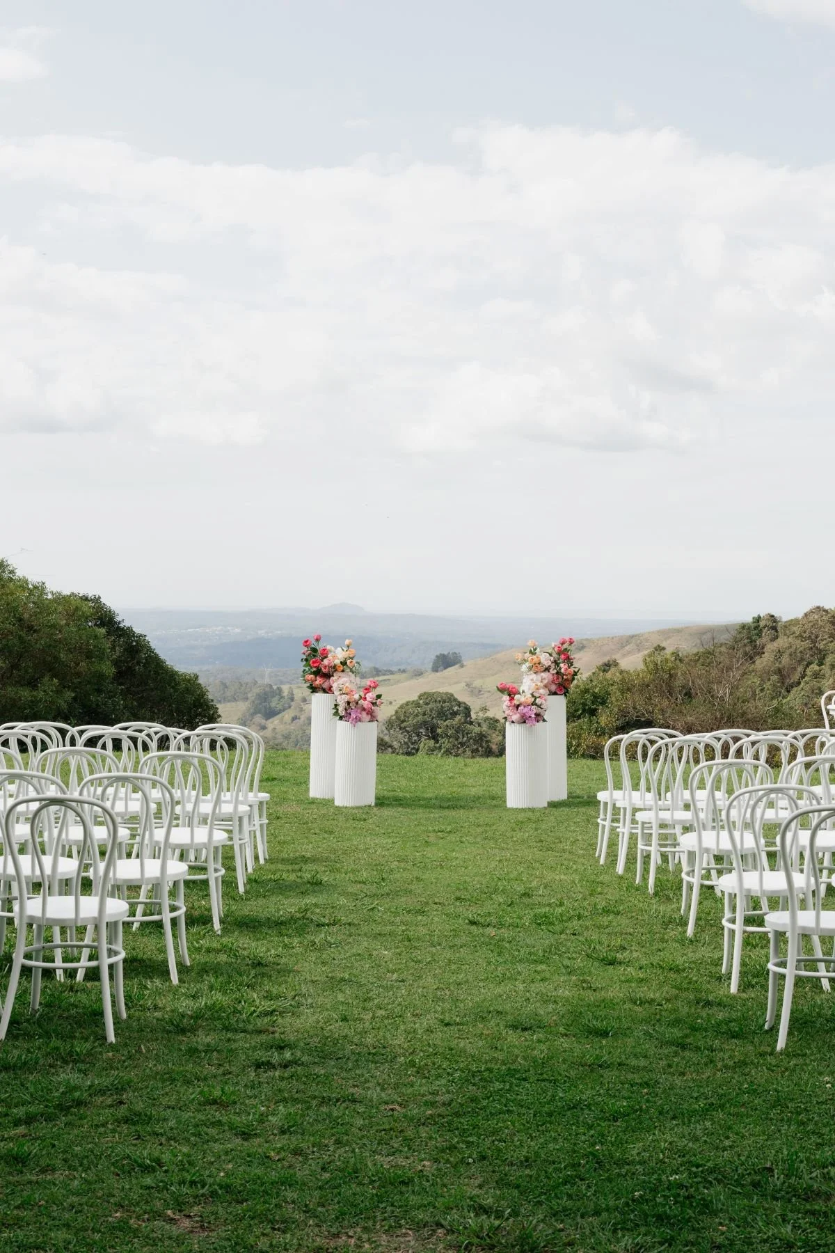 Outdoor wedding ceremony setup on Maleny Manor's Helipad with white bentwood chairs on each side of a grassy aisle, floral arrangements on tall white pedestals at the end of the aisle, with a scenic landscape of the Sunshine Coast.