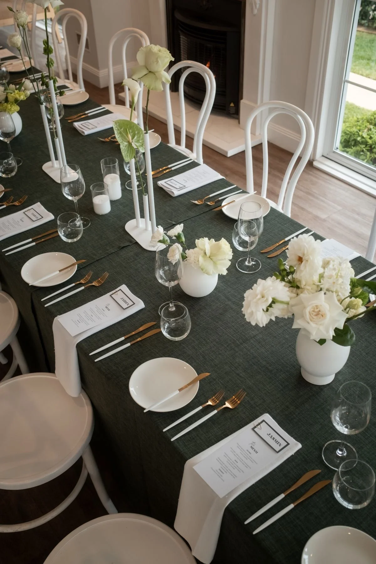 A decorated dining table set for a wedding with white flowers in vases, candles, wine glasses, and gold and white cutlery, with a dark green tablecloth and white bentwood chairs.