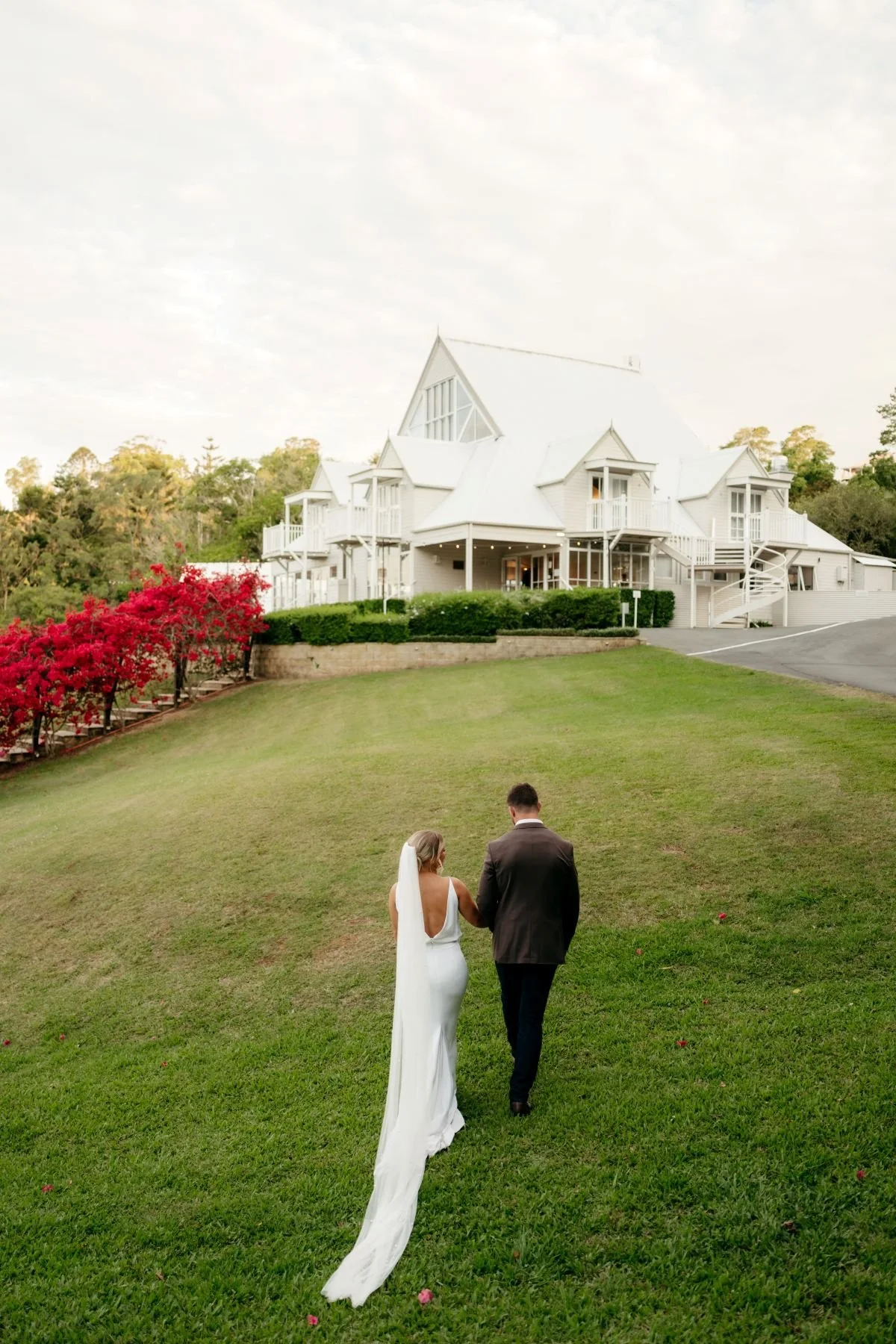 A bride and groom walking on a lush green lawn toward the Manor with multiple porches and balconies, surrounded by vibrant pink bougainvillea and trees.