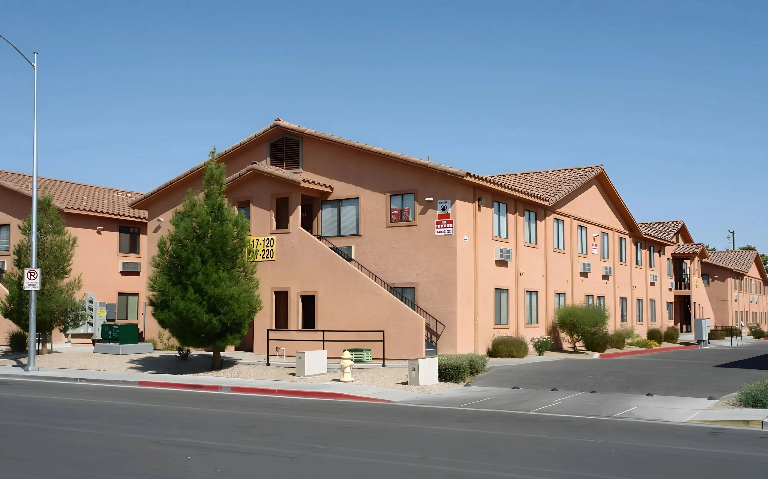 A multi-story apartment complex with peach-colored walls and tiled roofs, surrounded by trees, bushes, and parking spaces, under a clear blue sky.
