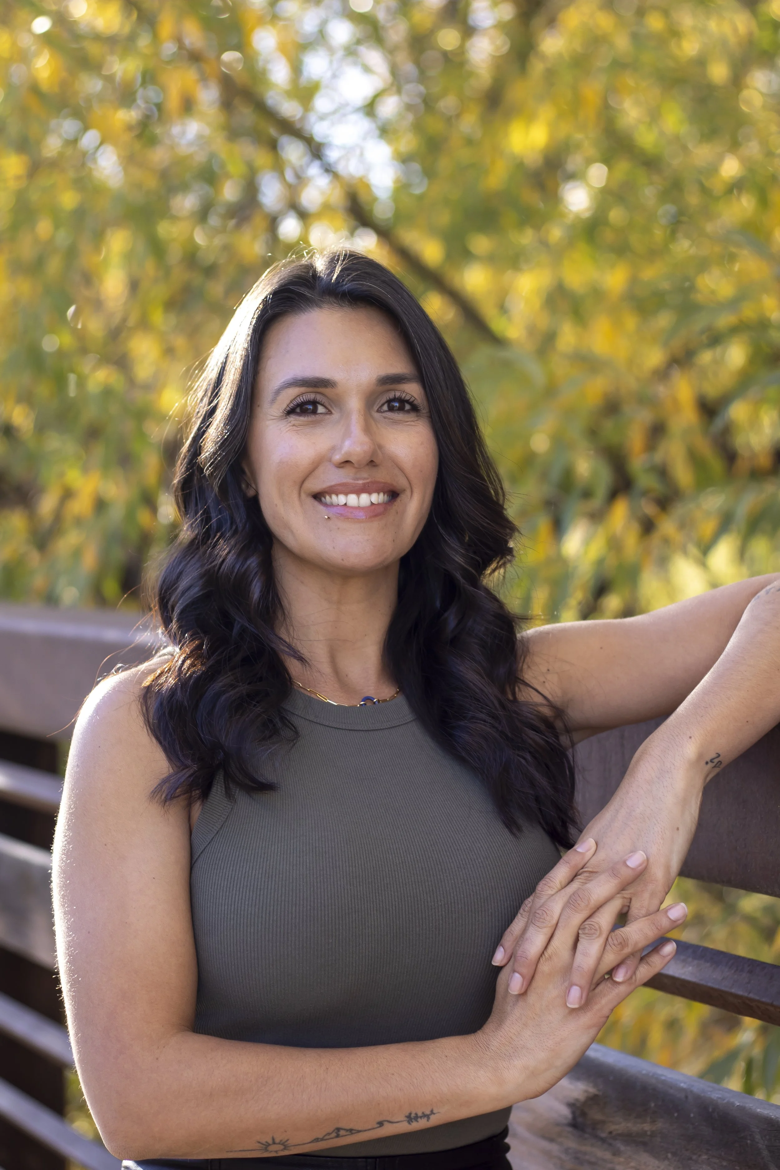 A woman with dark wavy hair smiling, wearing a sleeveless olive green top, leaning on a wooden railing outdoors with fall-colored trees in the background.