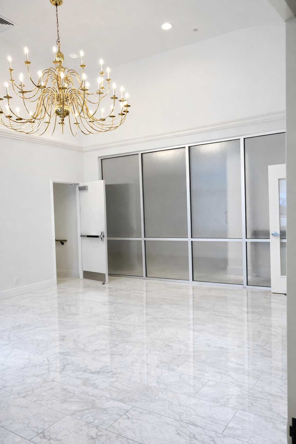 Empty room with white walls, marble floor, glass partition, and a large gold chandelier hanging from the ceiling.