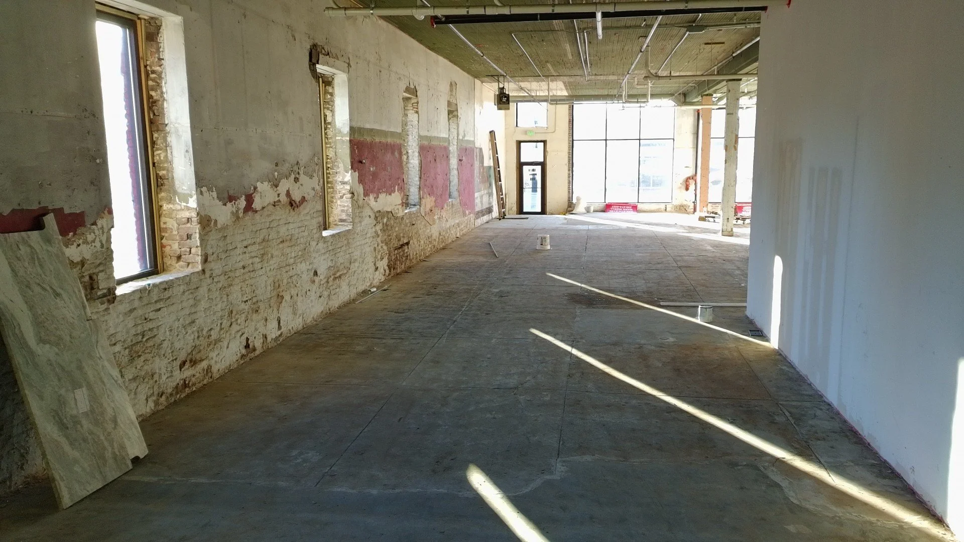 Interior of a building under renovation with exposed brick walls, large windows, and construction materials on the floor.