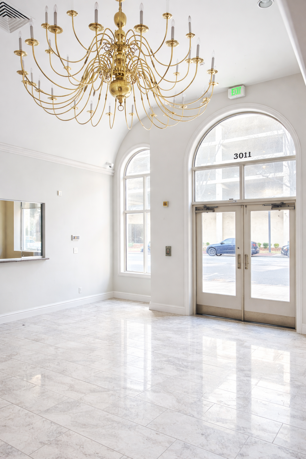 Bright and empty lobby with white walls, large arched windows, a gold chandelier, and glass double doors marked with the number 3011.