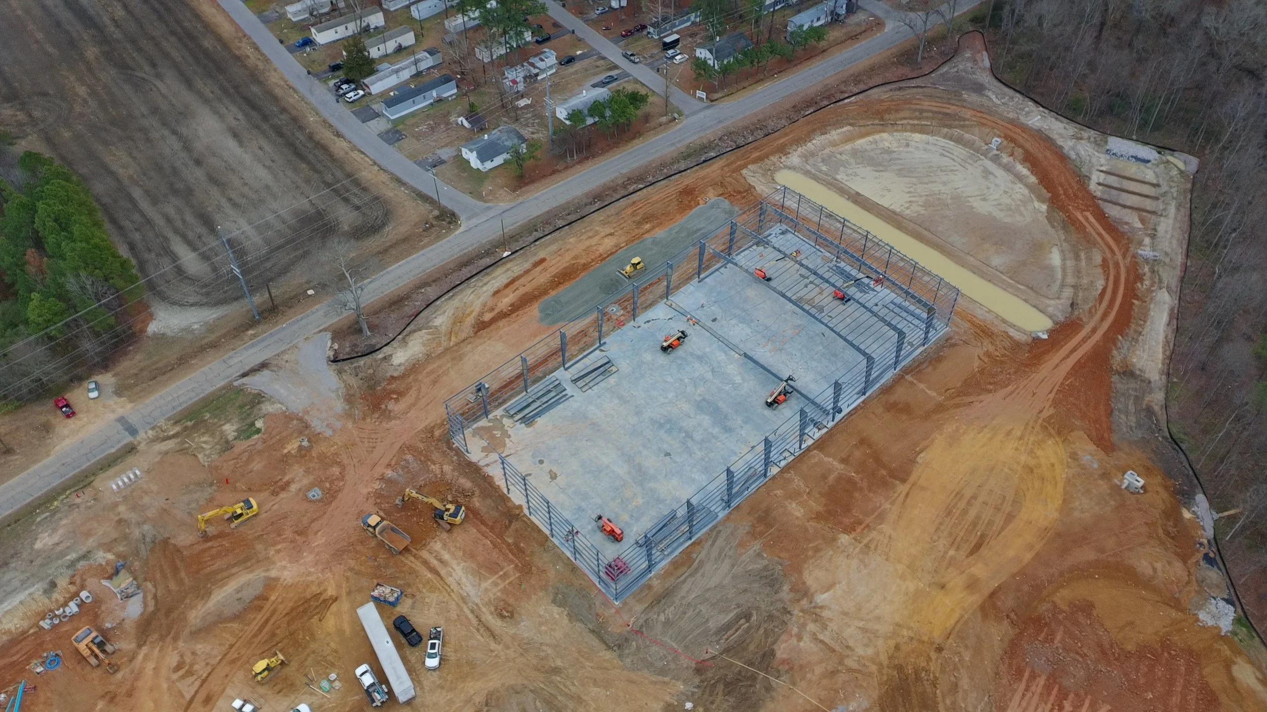 An aerial view of a construction site with a steel building framework being assembled. Surrounding the site are dirt roads and some construction vehicles, with a residential area nearby.