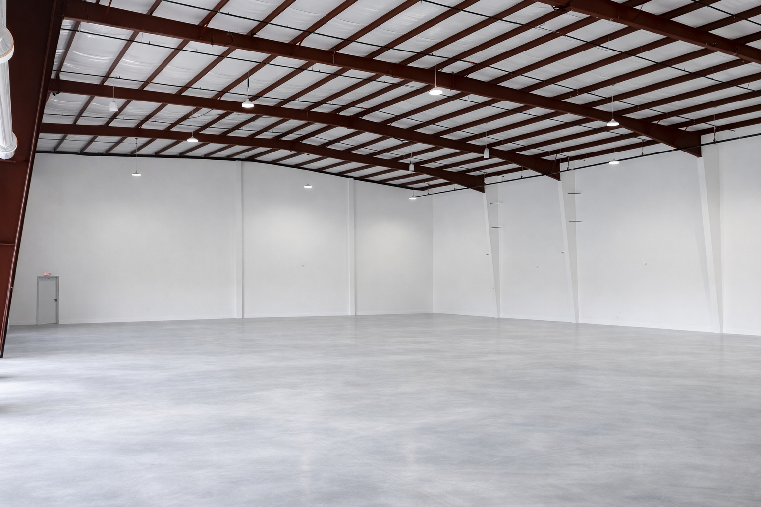 Empty indoor warehouse with white walls, high ceiling with exposed red beams, and a polished concrete floor.
