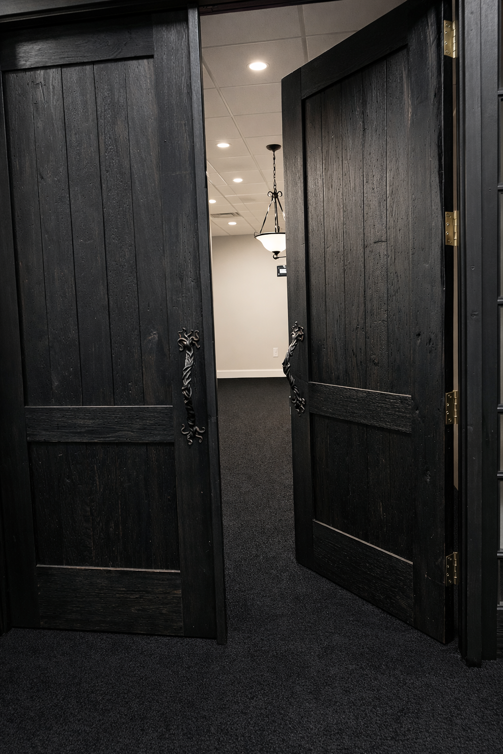 Open black wooden double doors with decorative handles, leading into a room with a white ceiling, hanging light fixtures, and dark carpeted floor.