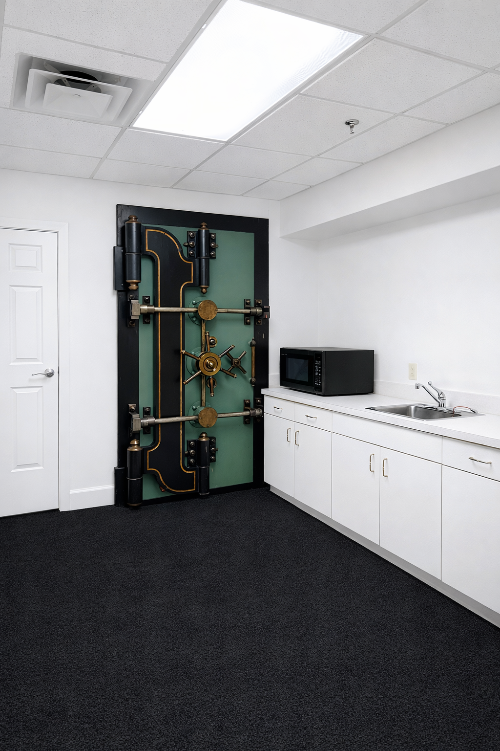A door with a large, decorative bank vault door in a white room with a microwave, sink, and cabinets.