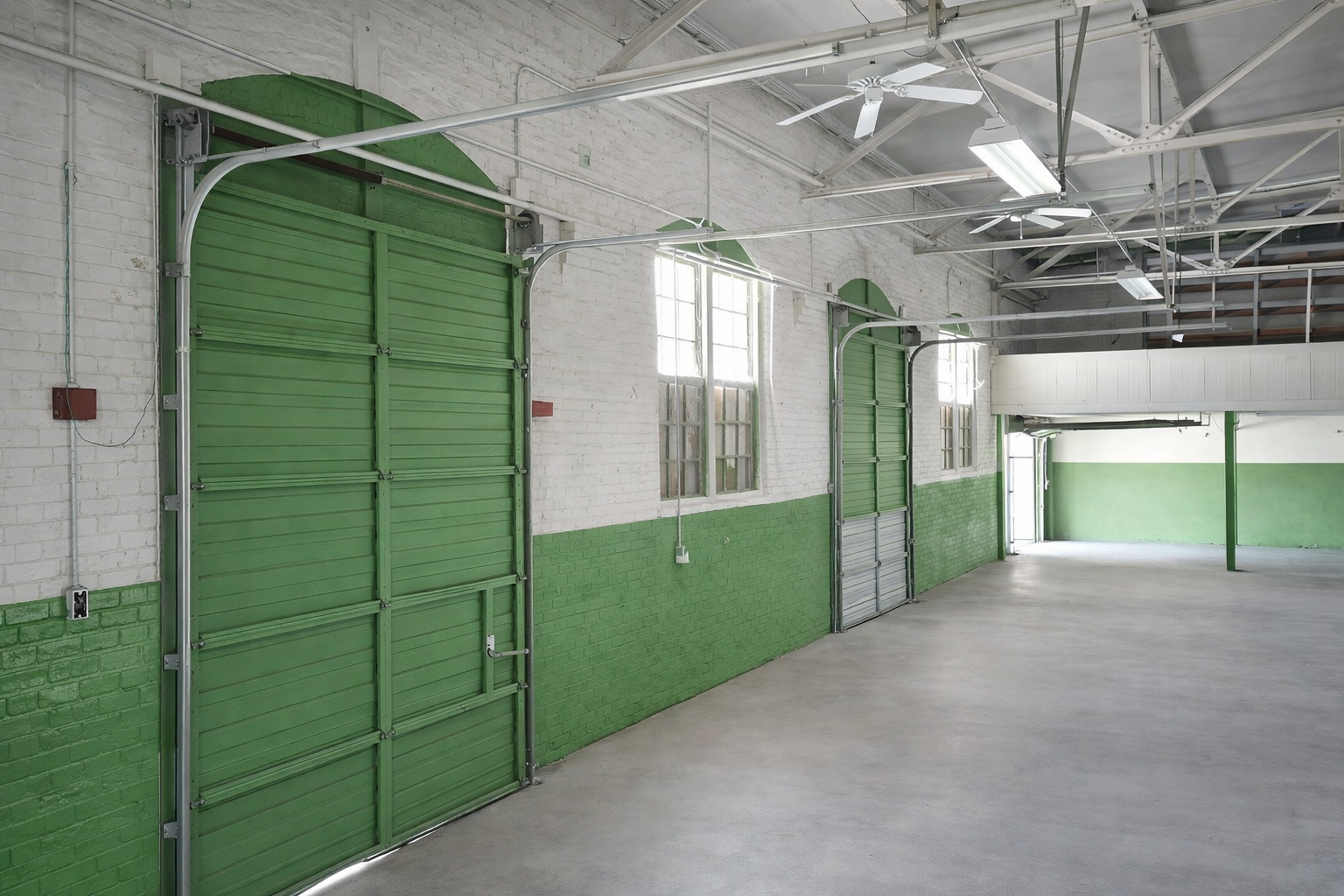 Empty industrial warehouse with white brick walls, green accents, multiple green roll-up doors, windows, ceiling fans, and fluorescent lights.