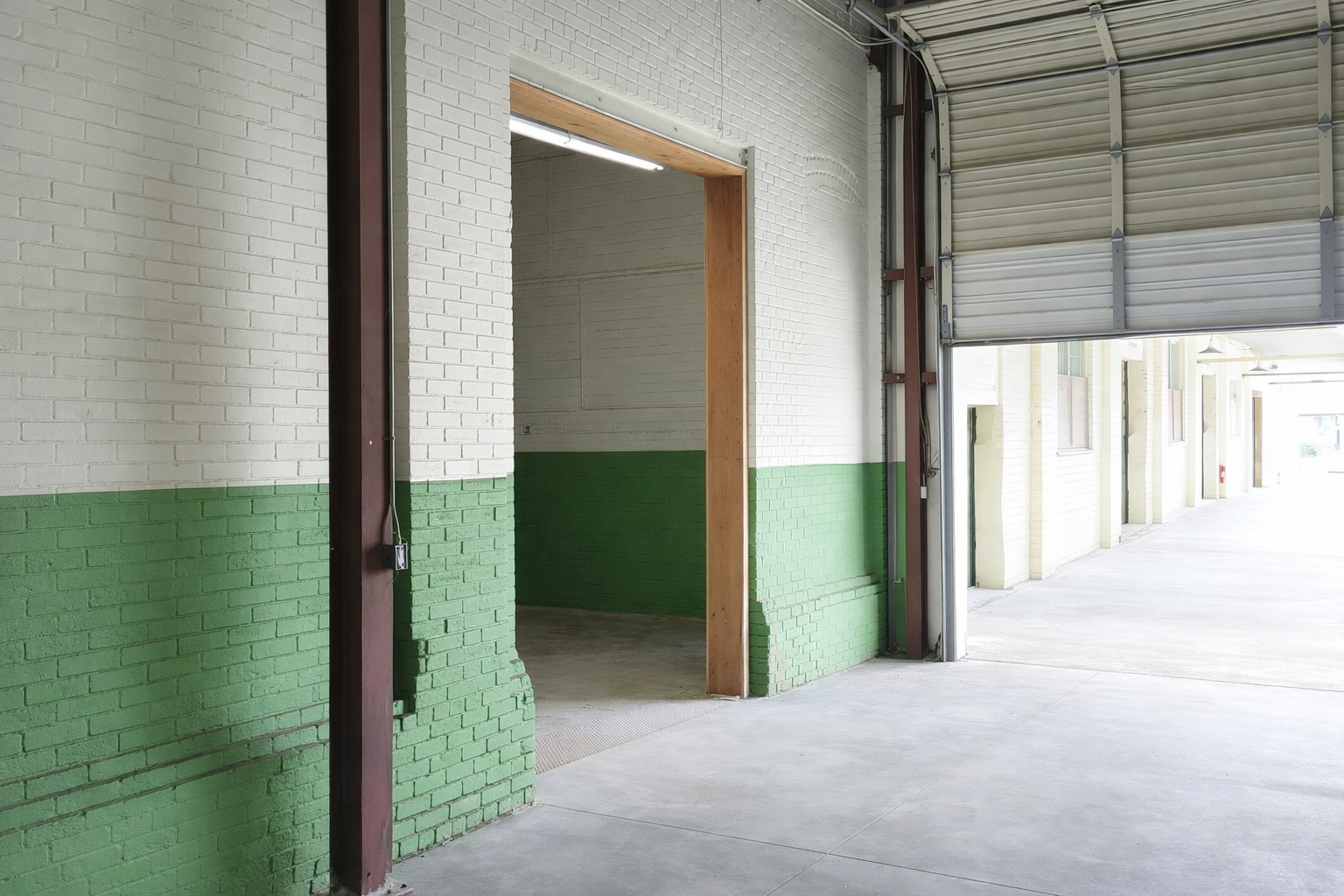 An empty warehouse or garage with white and green brick walls, a concrete floor, and a partially open roll-up garage door revealing a bright outdoor area.