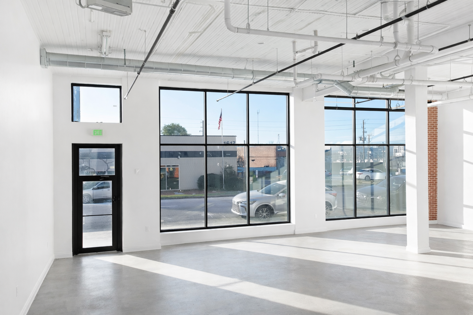 Empty commercial space with large front windows, a black door, exposed ceiling pipes, and a brick accent wall.