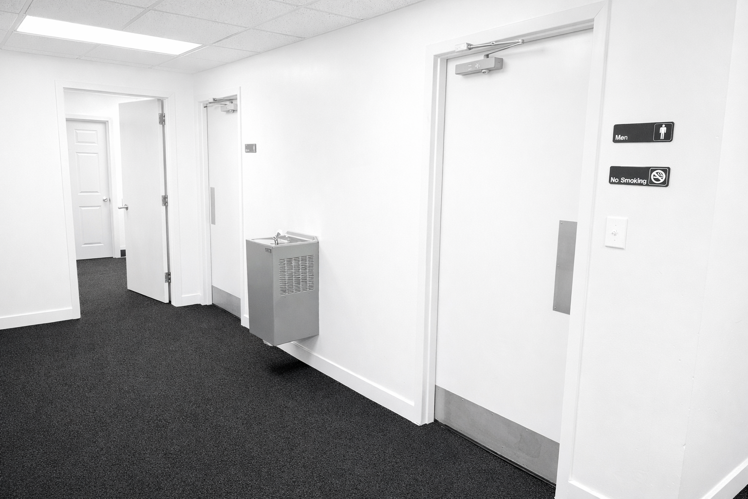 A hallway with white walls, dark carpeted floor, and multiple doors, including a men's restroom door with signage, a no smoking sign, and an accessible door closer.